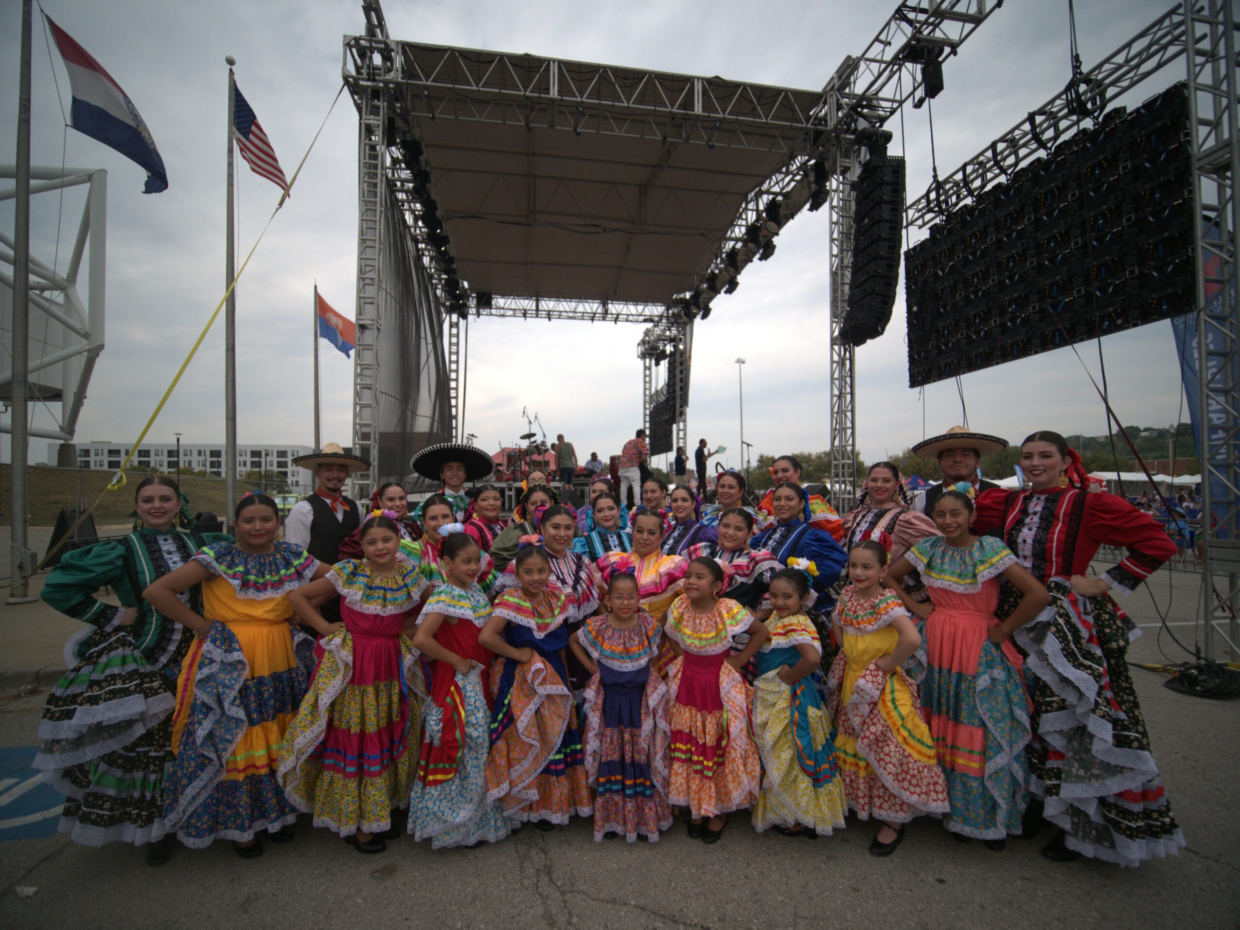 Group of people in colorful traditional dresses posing on stage outdoors with flags and stage equipment behind them.