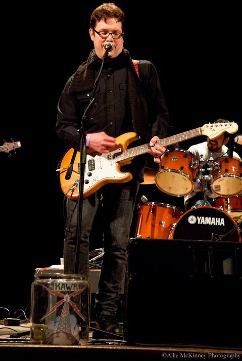 Person playing electric guitar and singing into microphone on stage with drums in background.