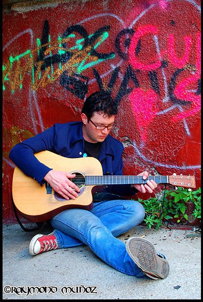 Person sitting on ground playing an acoustic guitar against a graffiti-covered wall.
