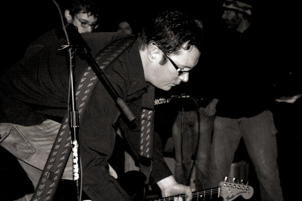 Person with glasses playing guitar on stage, with microphone stand nearby, in a dark setting.