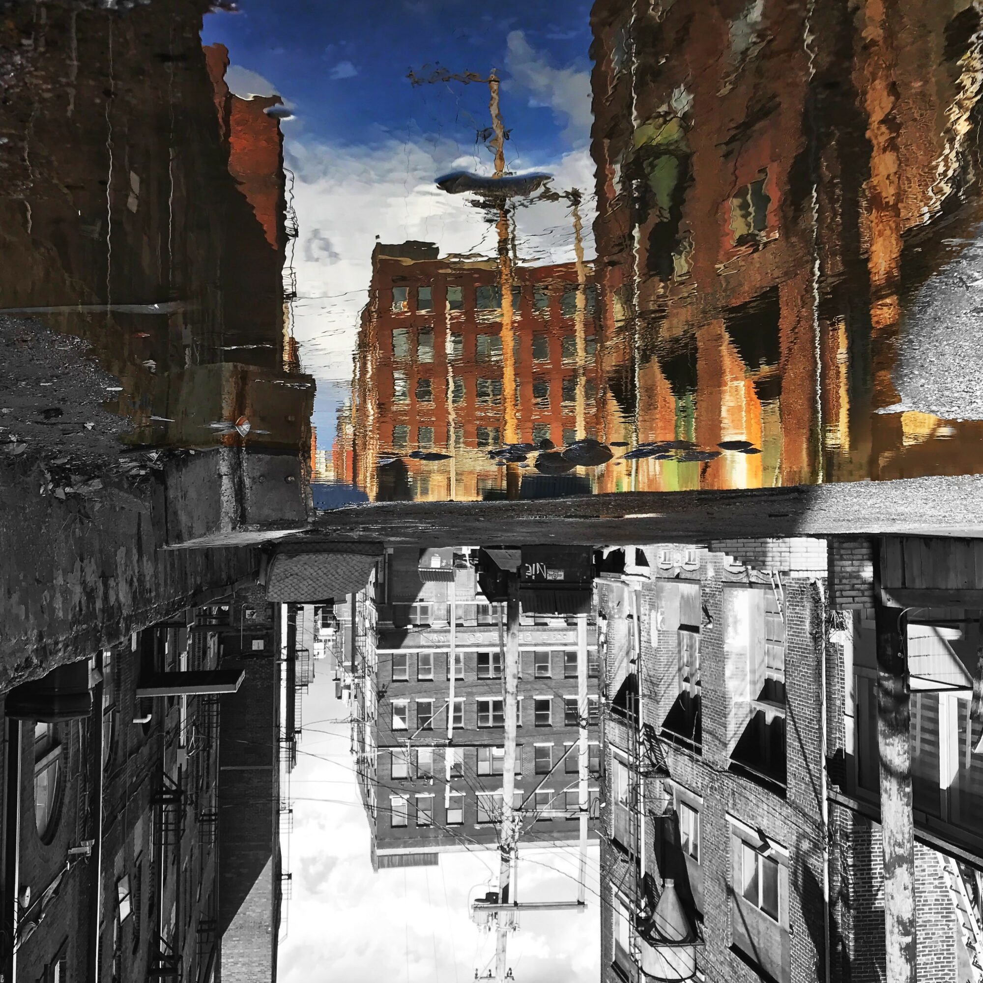Upside-down city street with buildings, a fountain, and a cloudy sky, reflected in a puddle on the ground.