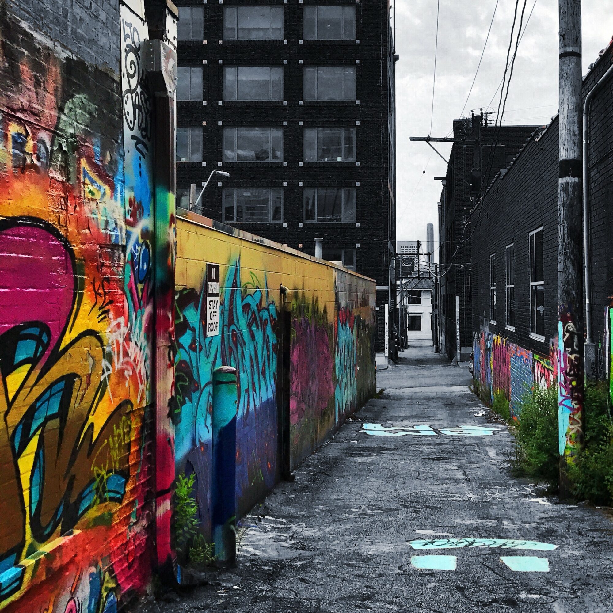 Urban alleyway with graffiti-covered walls, leading to a building in the distance, overcast sky, wet pavement, and power lines.