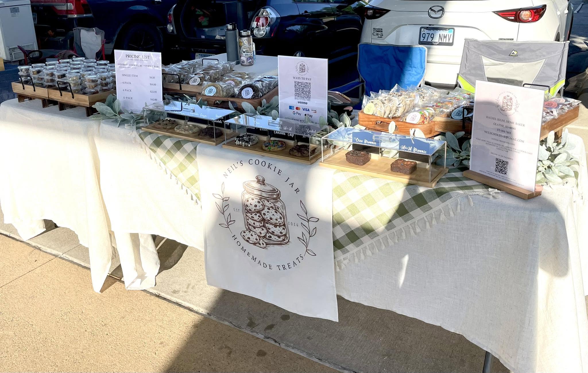 Outdoor market stall with baked goods and signs on a table covered with a white cloth, cars parked behind.
