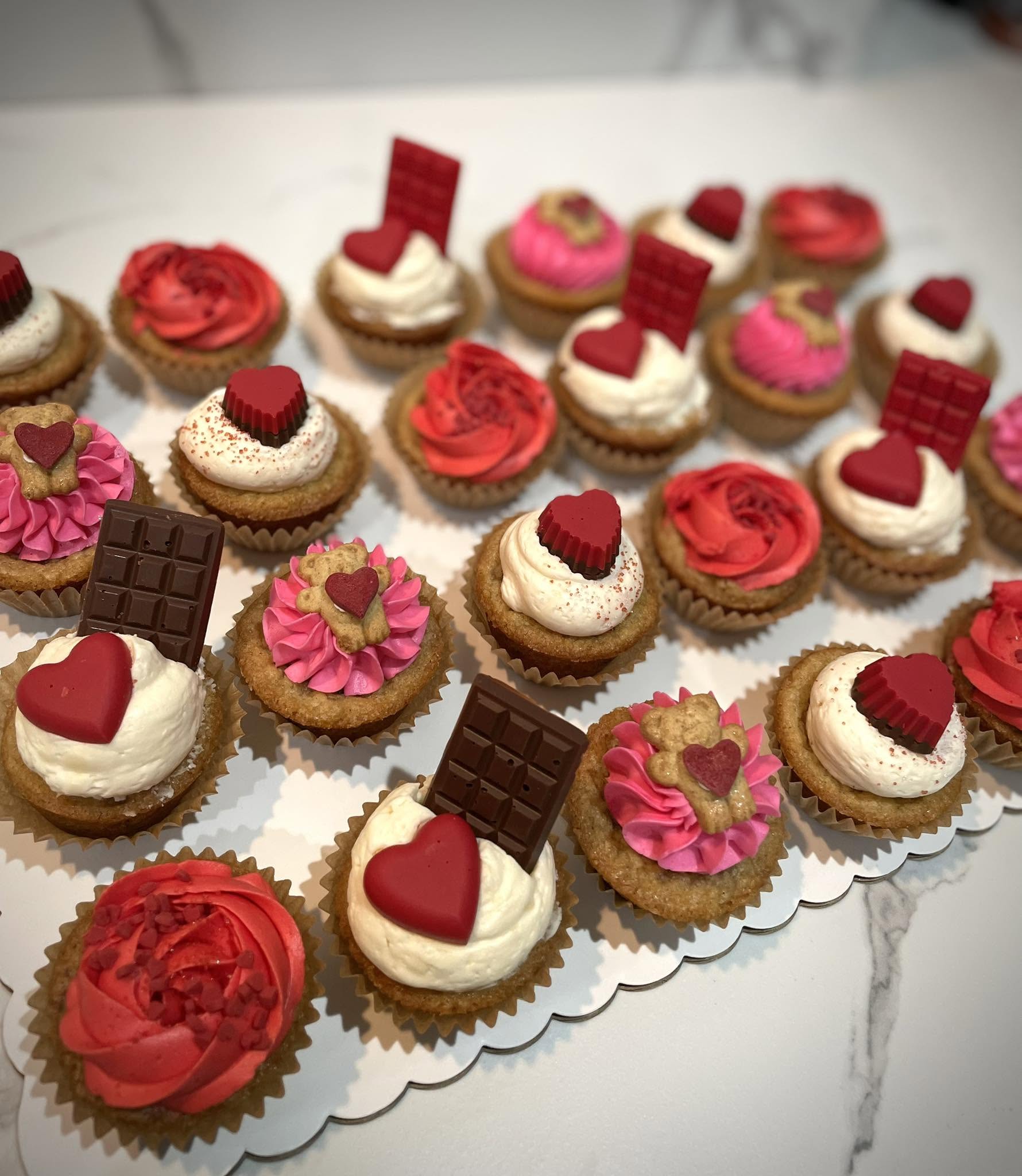 Assorted decorated cupcakes with pink, red, and white frosting, chocolate pieces, and heart-shaped toppings on a white surface.