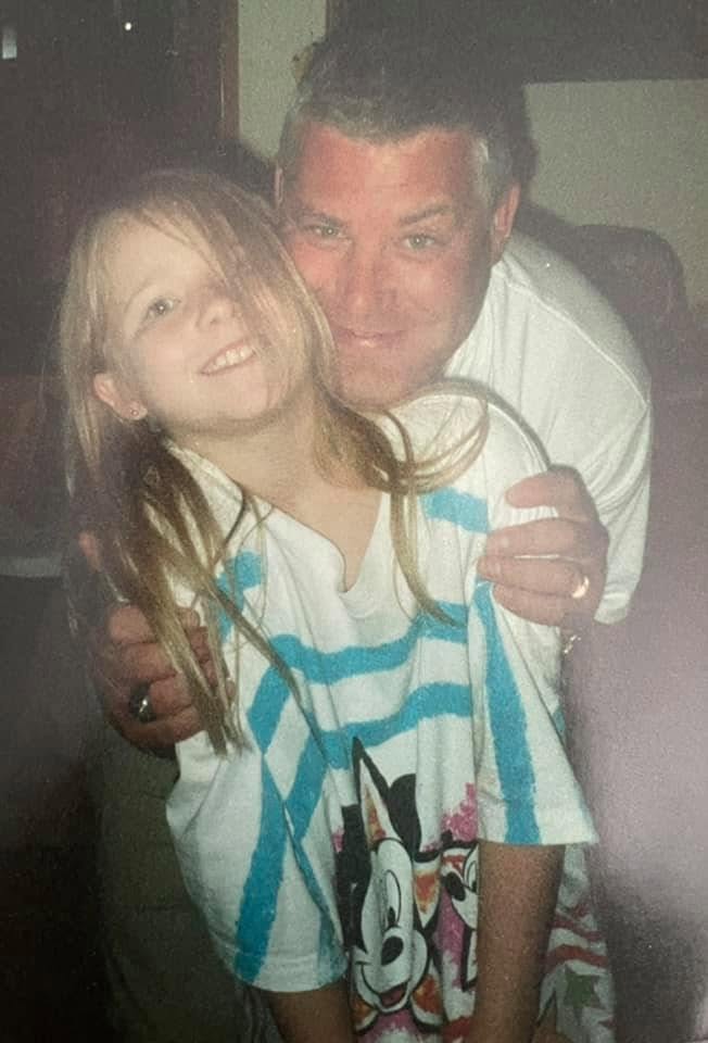 A man and a young girl are smiling and hugging indoors, the girl wearing a striped shirt with a cartoon character.