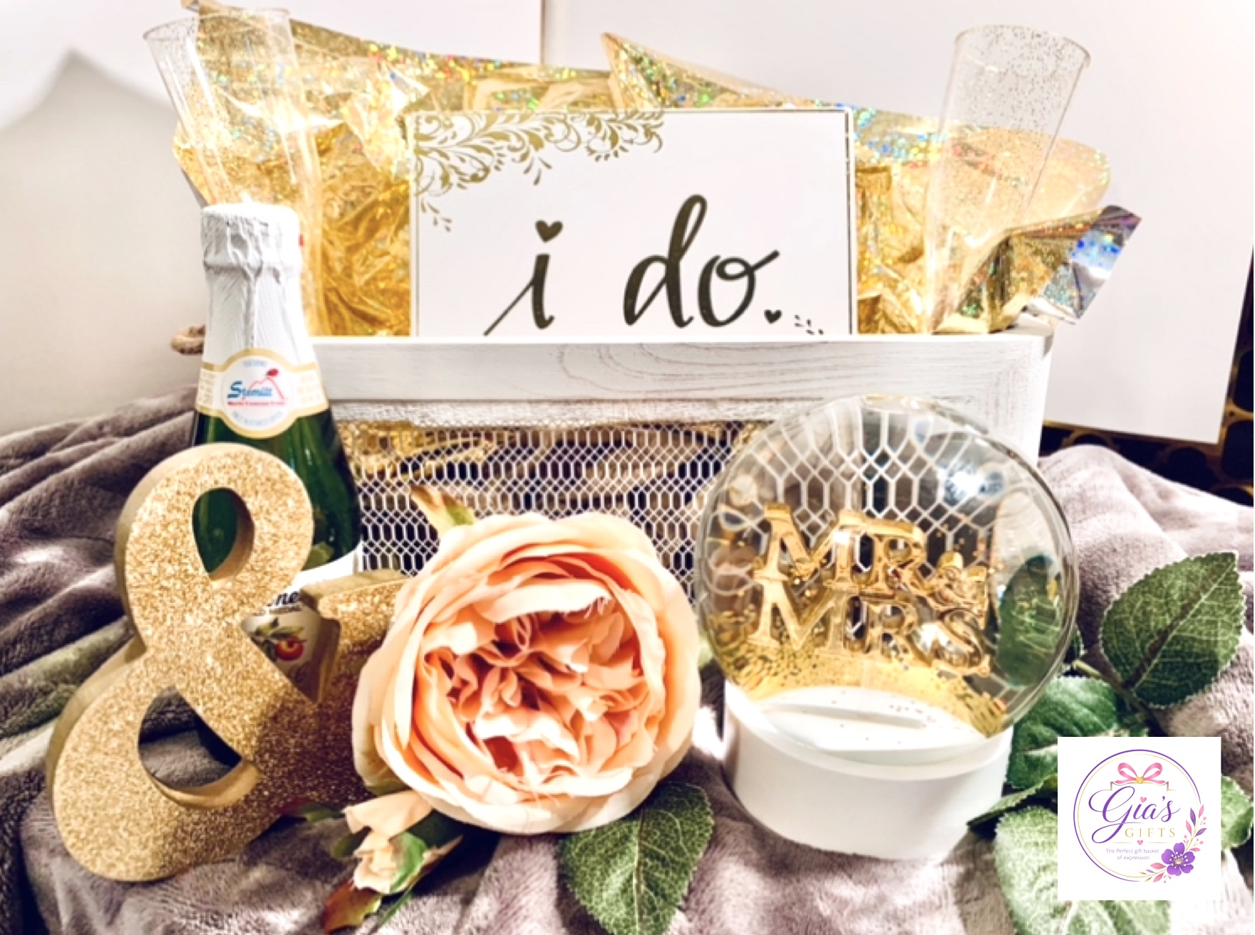 Decorative wedding or celebration setup with a sign reading 'i do', a large peach-colored flower, gold and glitter accents, and greenery.