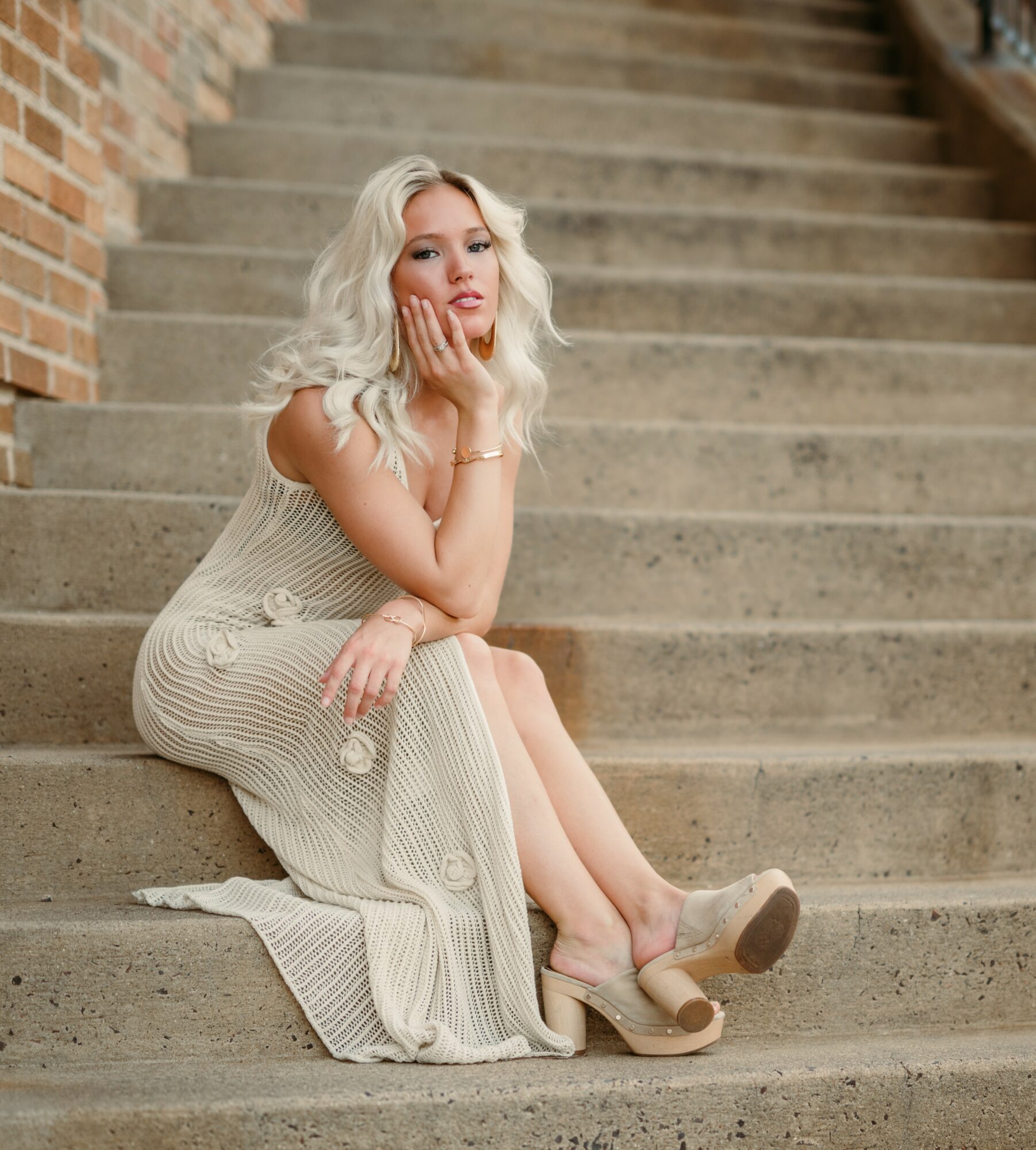 Woman with blonde hair sitting on stairs, wearing a beige dress and high heels, resting her chin on her hand.