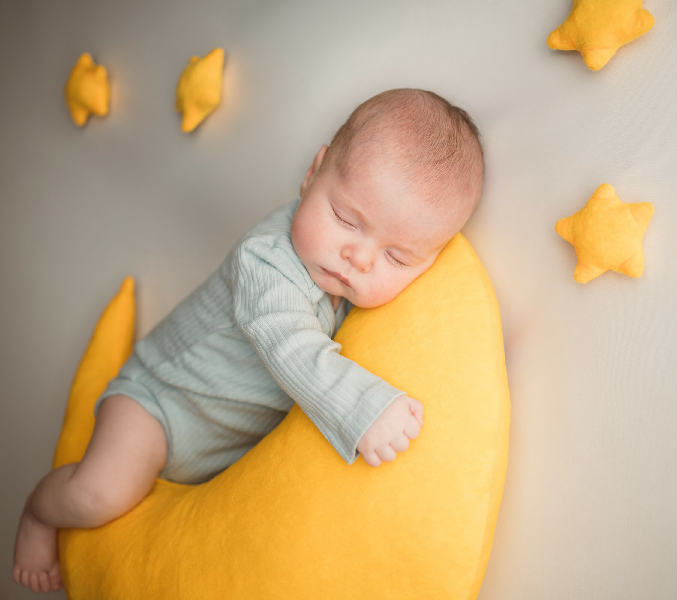 Baby sleeping on a yellow crescent-shaped pillow surrounded by yellow star-shaped plush toys, on a light background.