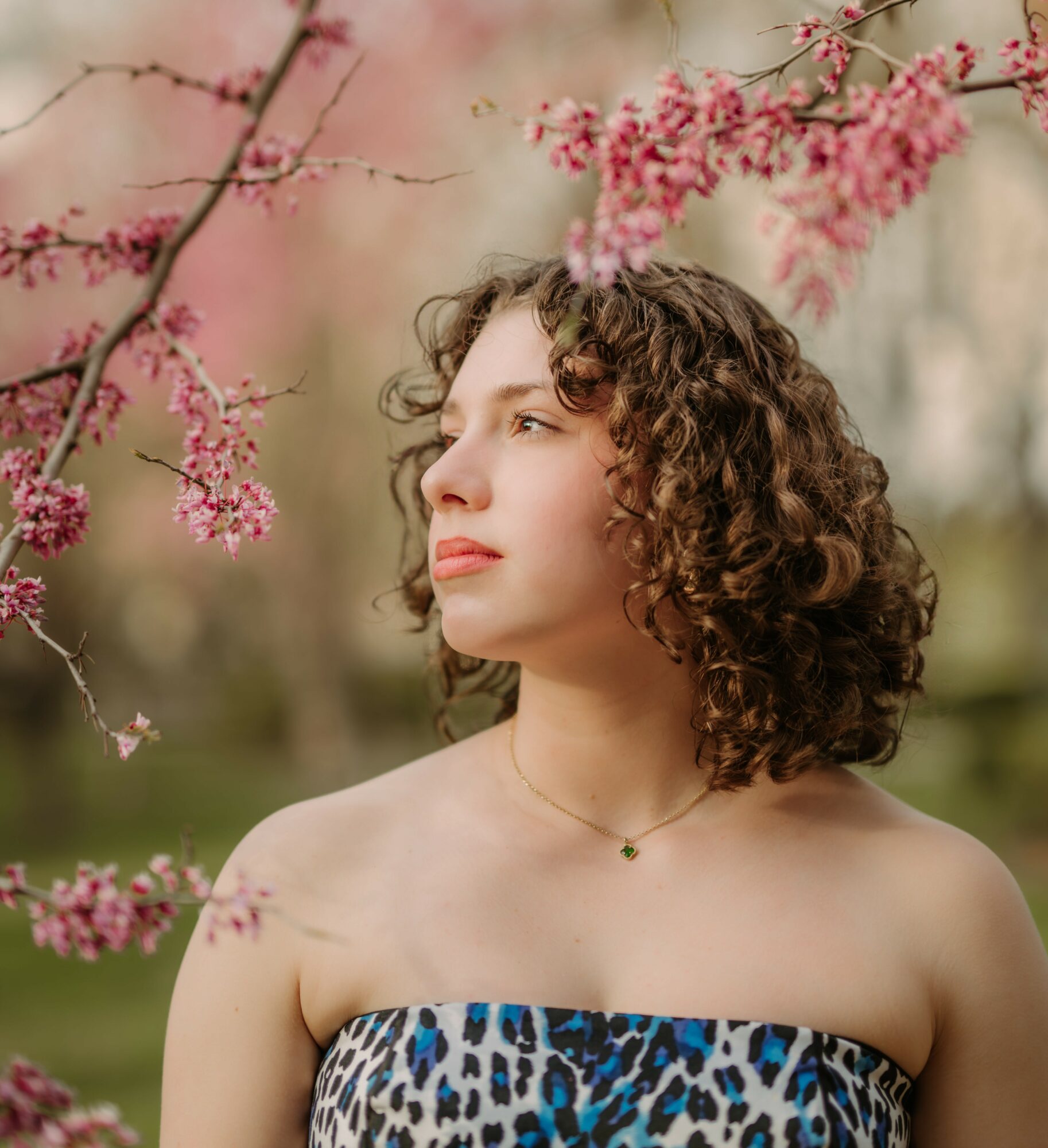 Young woman with curly hair looking to the side among pink blossoms outdoors.