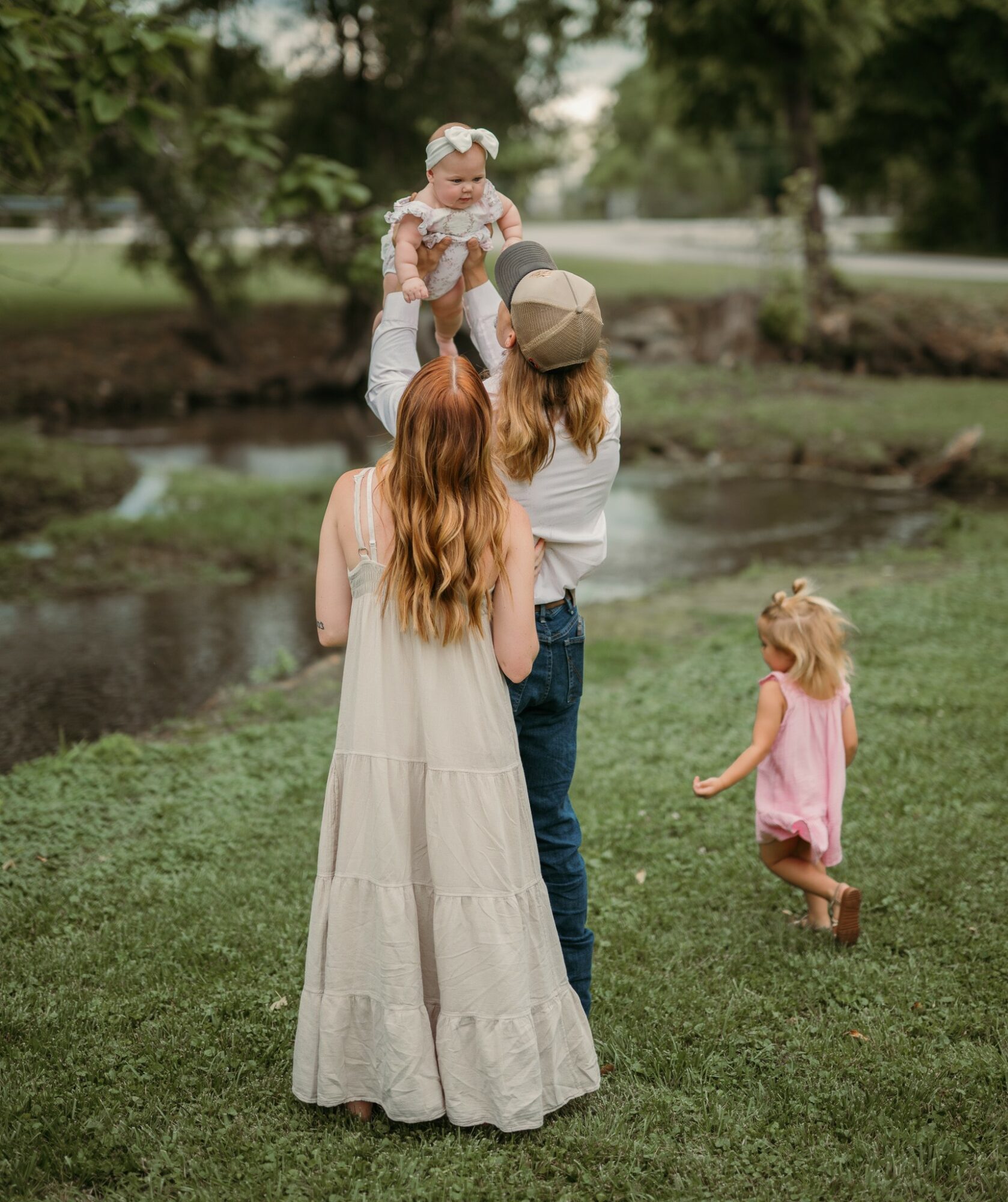 A woman holds a baby girl while two young girls stand nearby in a park with trees and a pond.