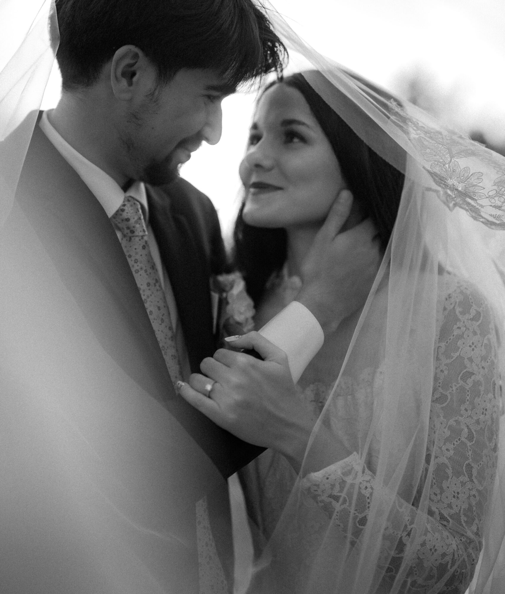 A bride and groom face each other closely, with the groom gently holding the bride's face, both smiling softly.