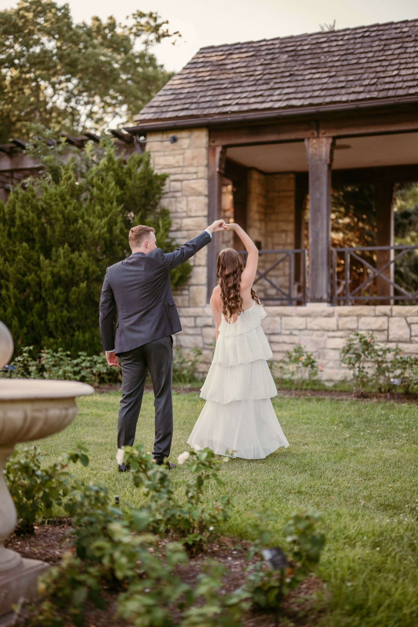 A couple dancing outdoors in front of a stone building with greenery, the man in a suit and the woman in a white dress.