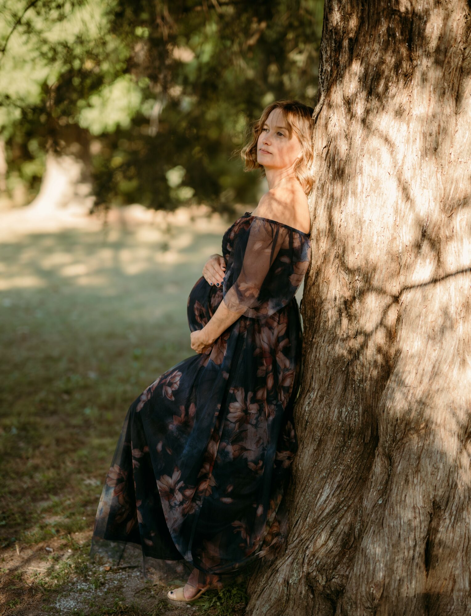 Woman in a dark floral dress leaning against a tree outdoors, sunlight filtering through leaves, serene expression.