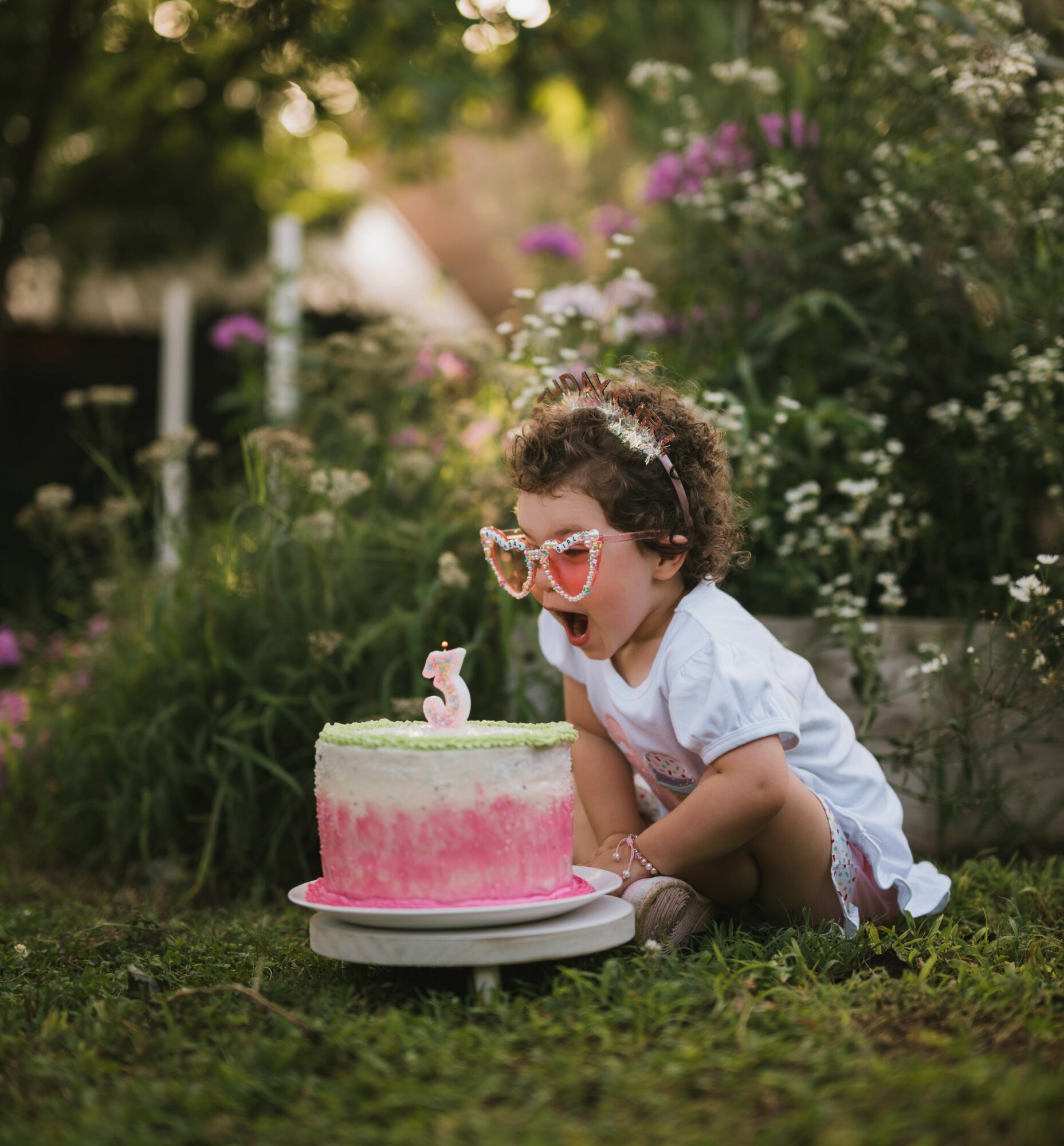 Child with curly hair and sunglasses celebrates birthday outdoors with pink cake and a number 3 candle.