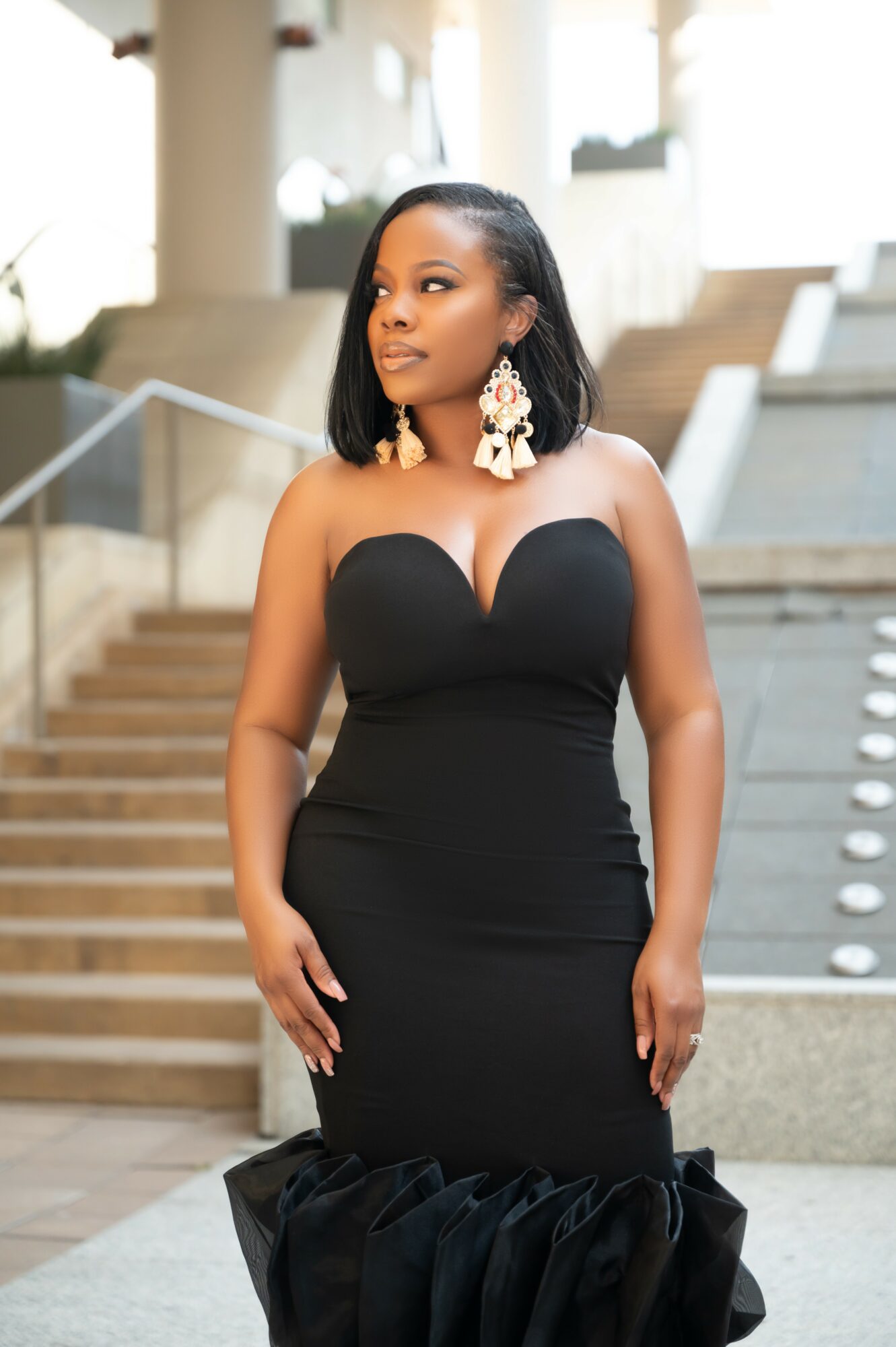 Woman in a strapless black dress with large earrings stands on stairs in a modern building.