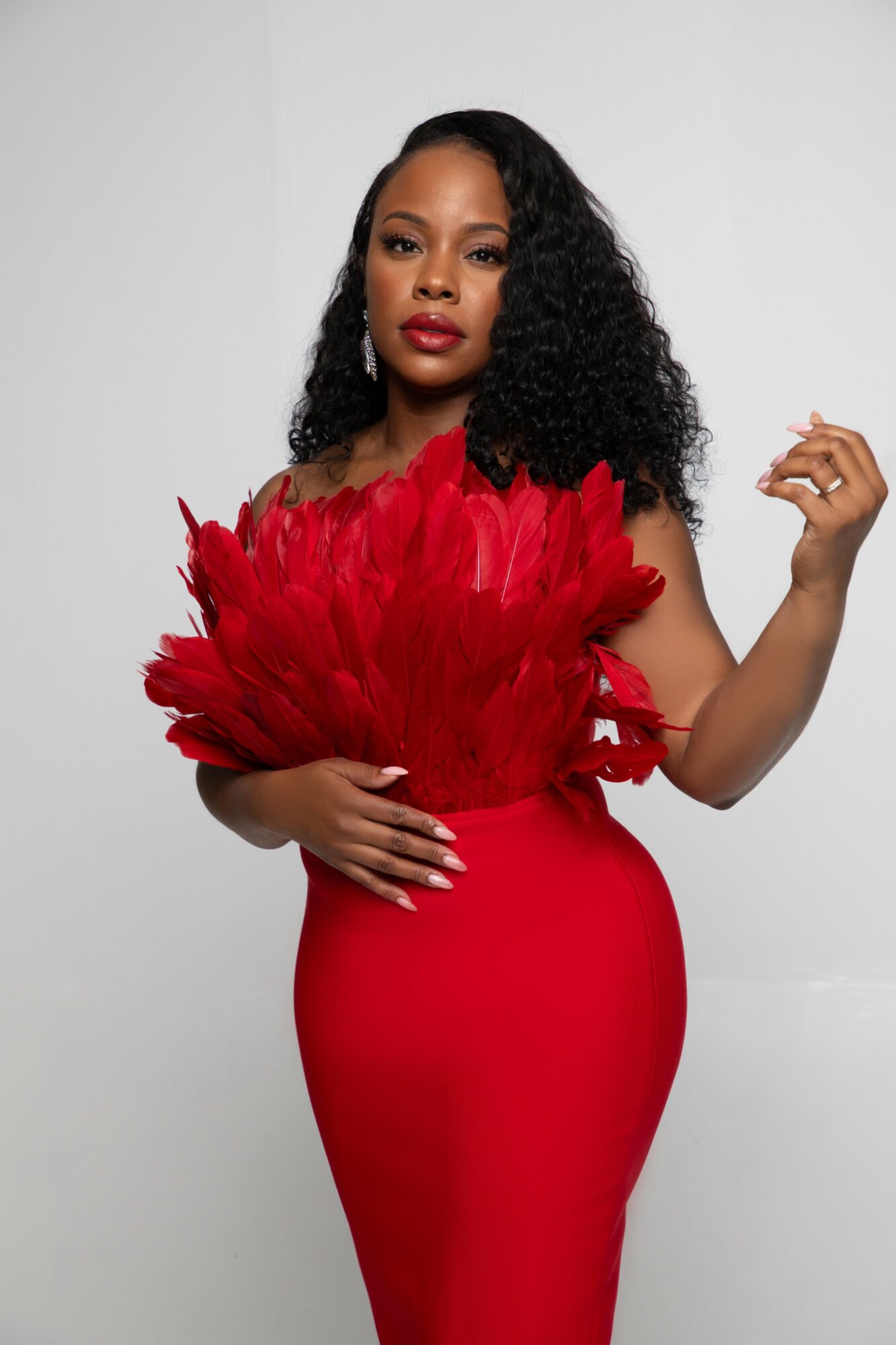 Woman in red dress holding red feathered accessory, standing against plain background.