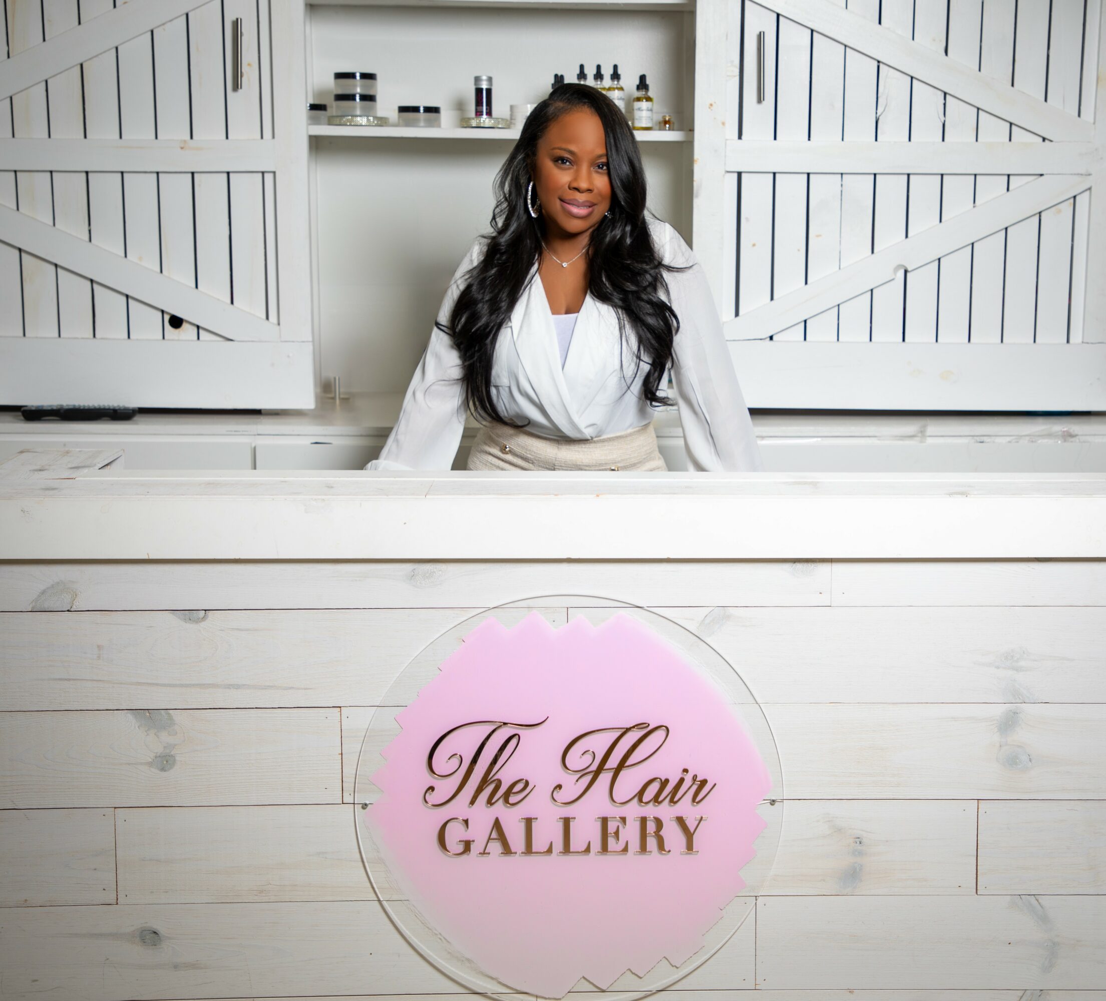 Woman standing behind a counter with a sign reading 'The Hair Gallery' in pink and black text.