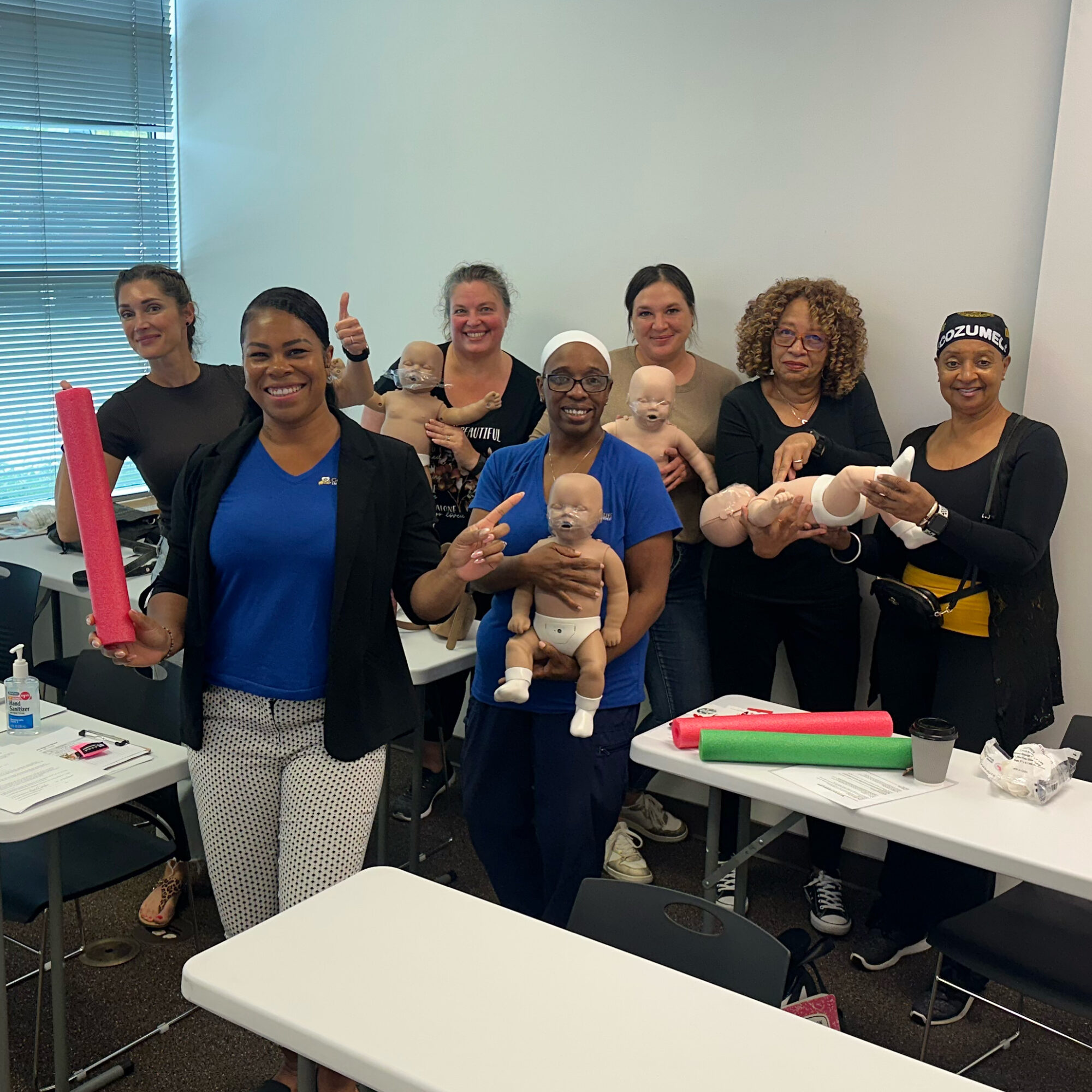 Group of seven women holding baby dolls in a classroom with tables and chairs, smiling and giving thumbs up.