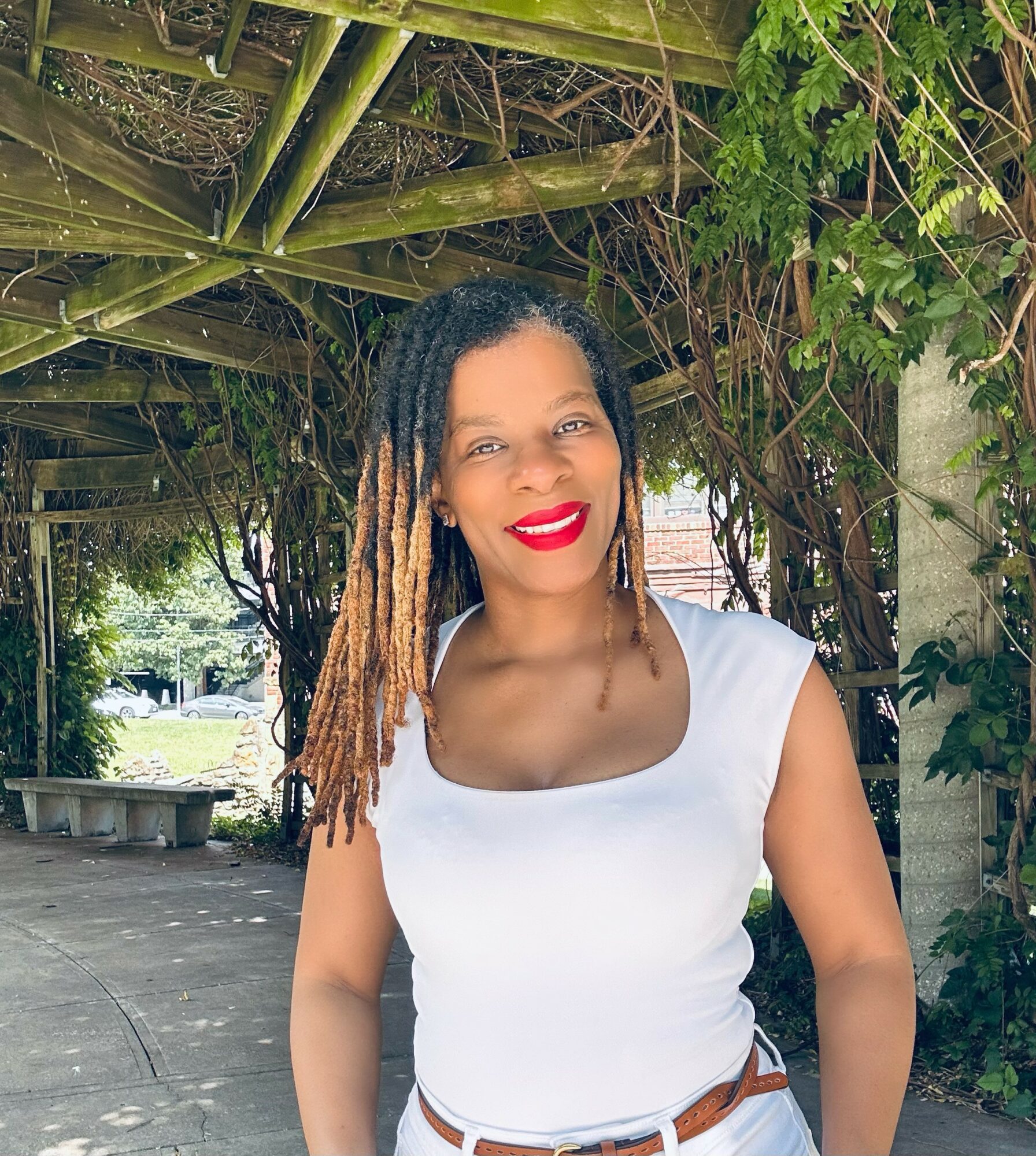 Woman with dreadlocks and red lipstick smiling outdoors under a wooden pergola, wearing a white top and brown belt.