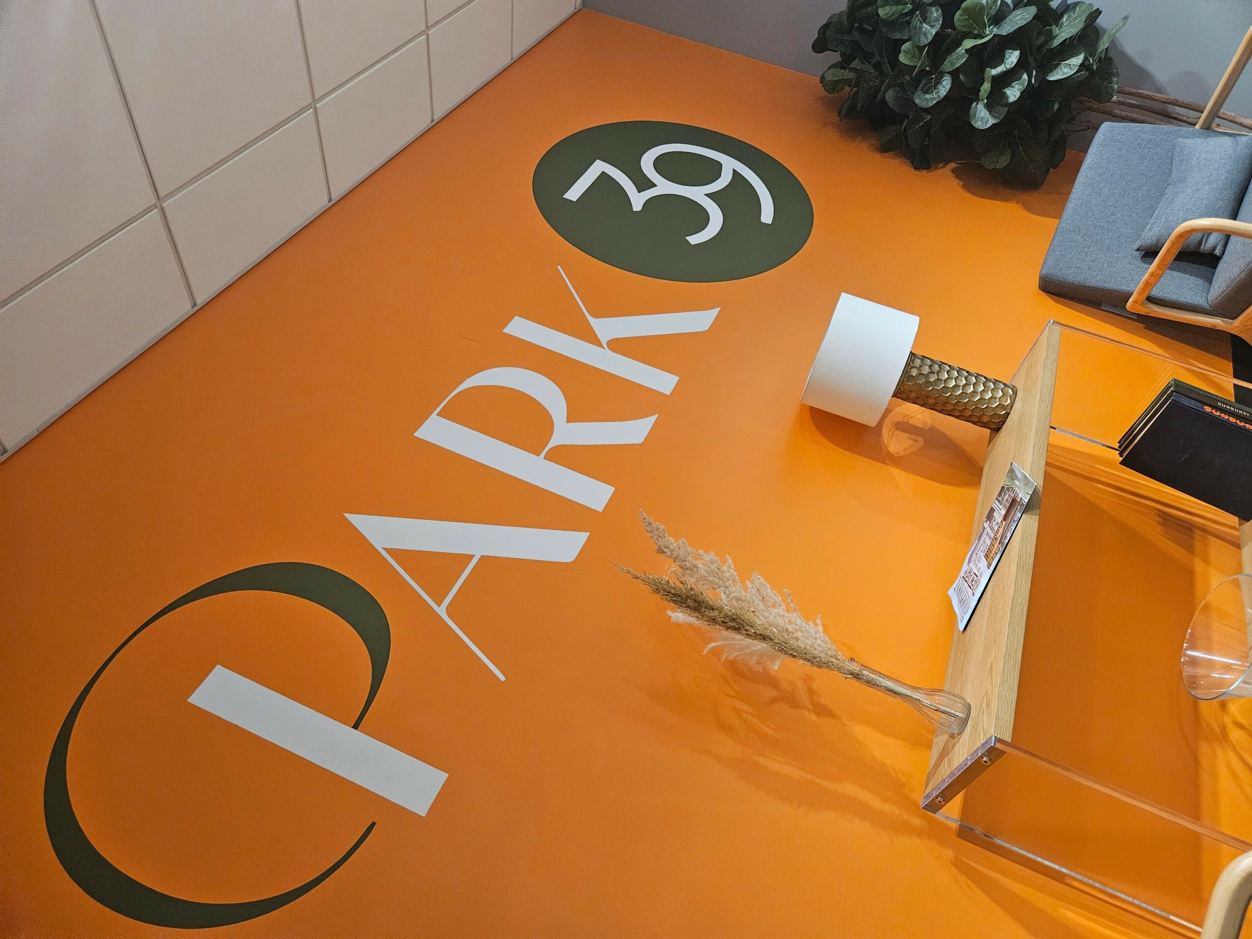 View of a desk with orange surface, a plant, a roll of tape, and a transparent container, with text and logo. Word: PARK.