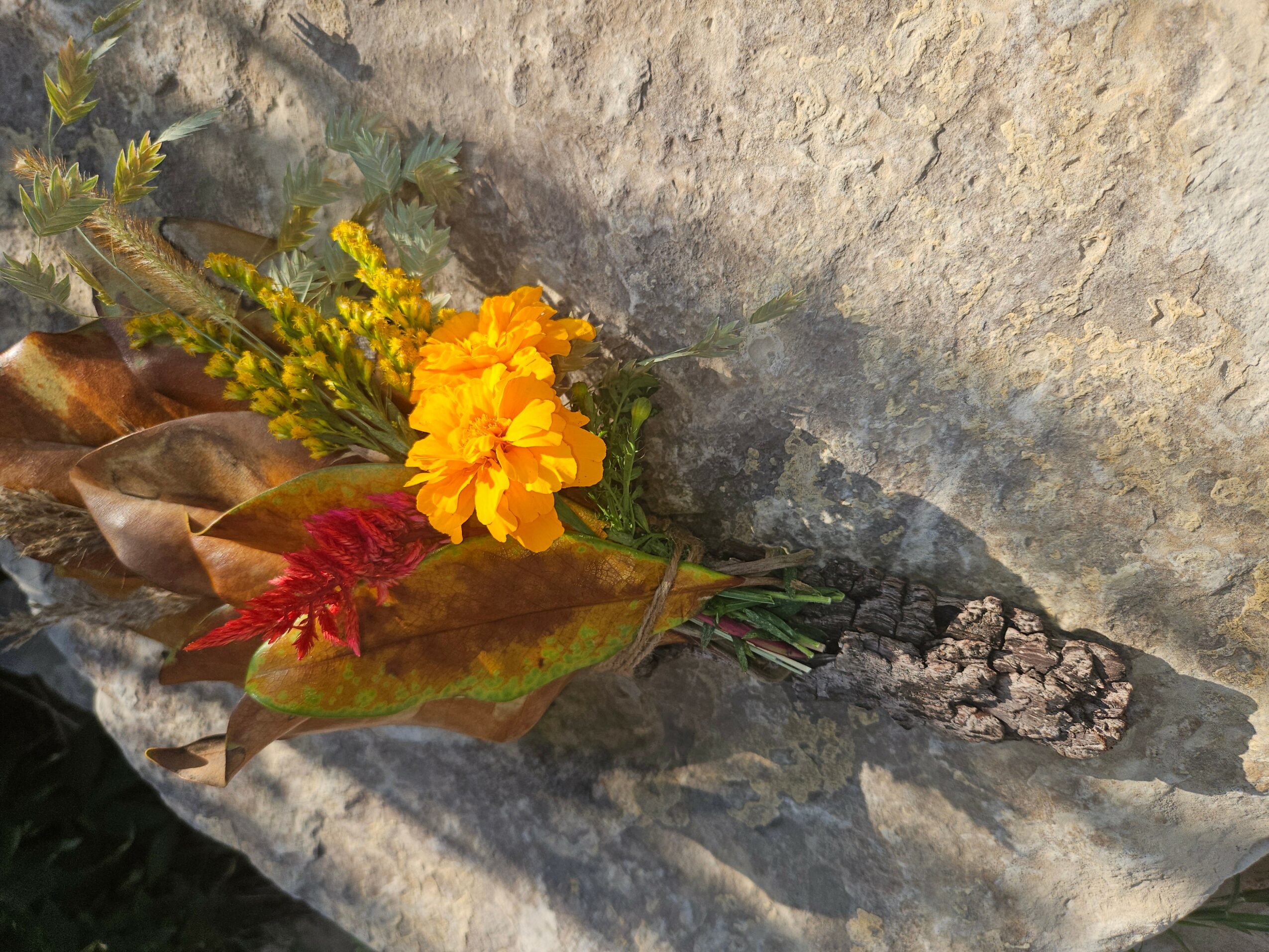A small bouquet of yellow flowers, red leaves, and dried brown leaves on a stone surface.