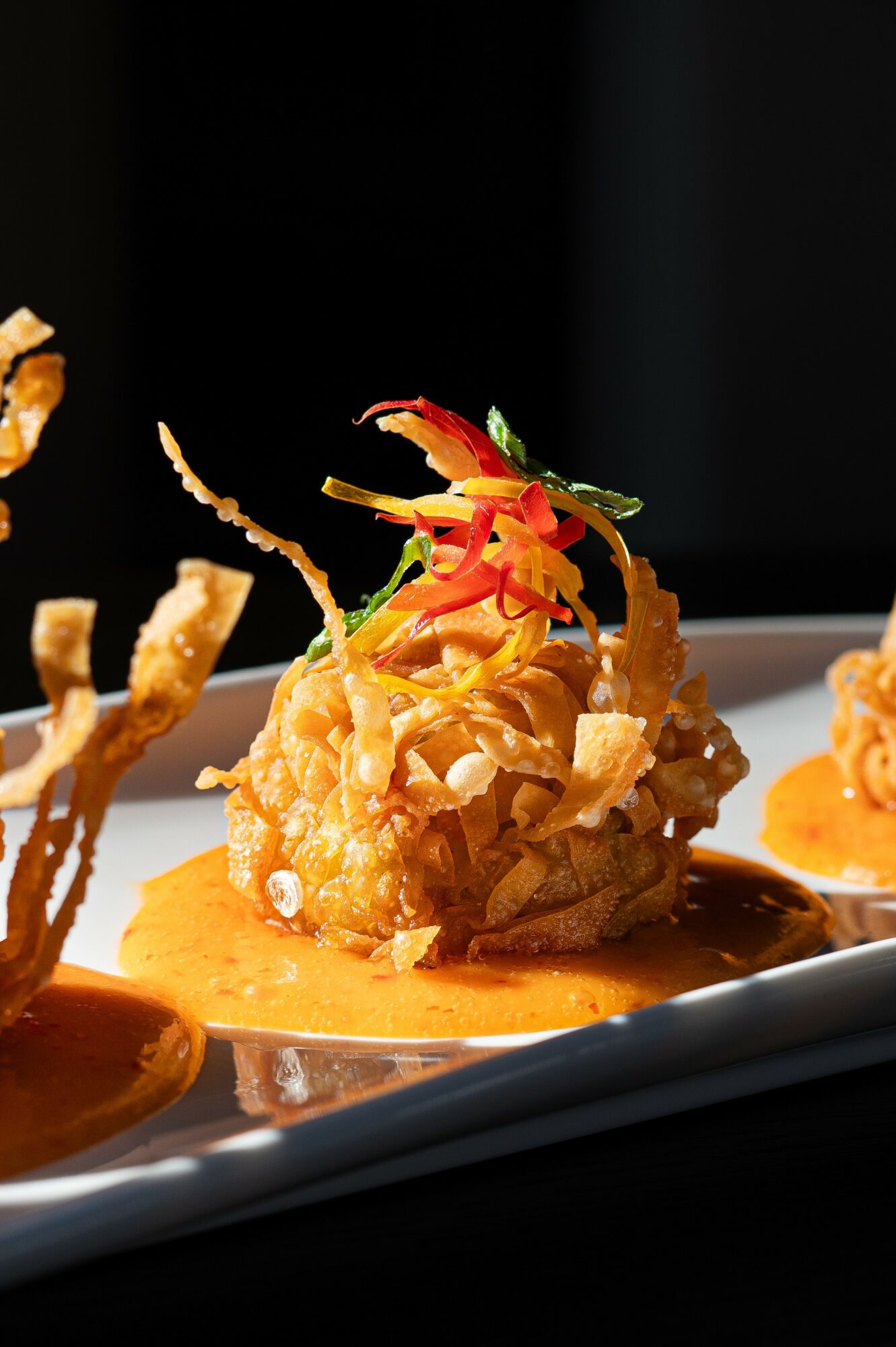 Fried appetizer topped with colorful vegetables and sauce on a black plate, with other similar dishes partially visible.