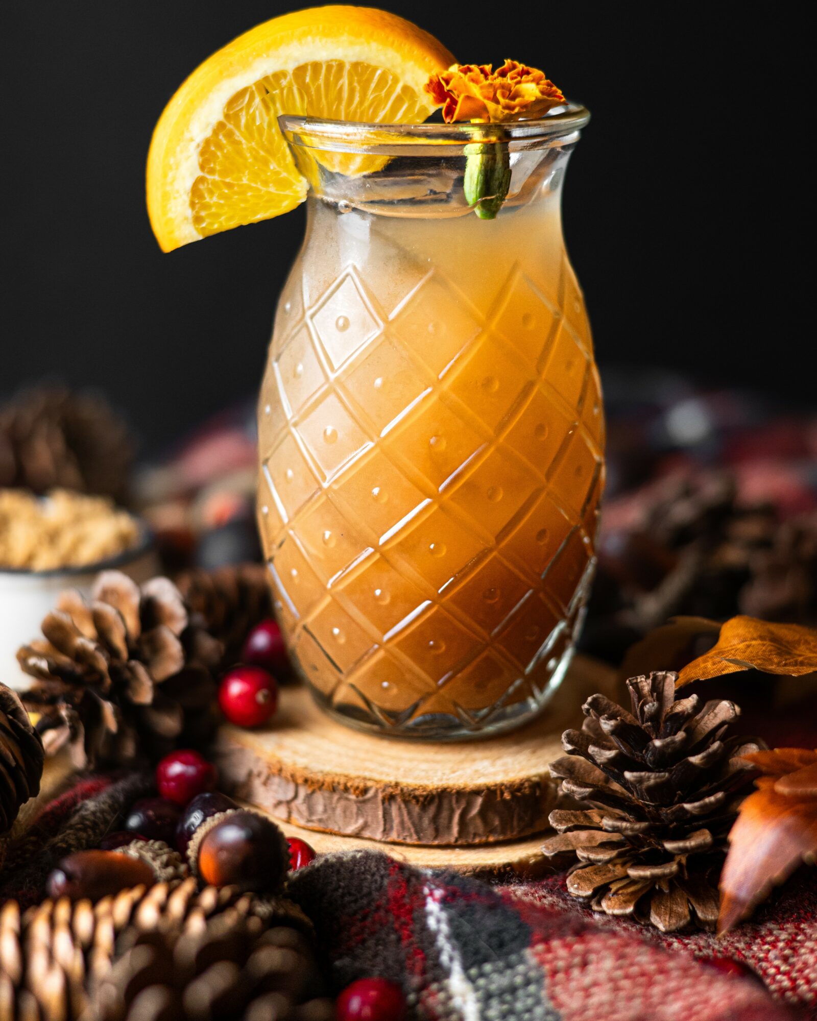 A glass of orange cocktail with lemon slice and garnish, surrounded by pinecones and berries on a wooden surface.