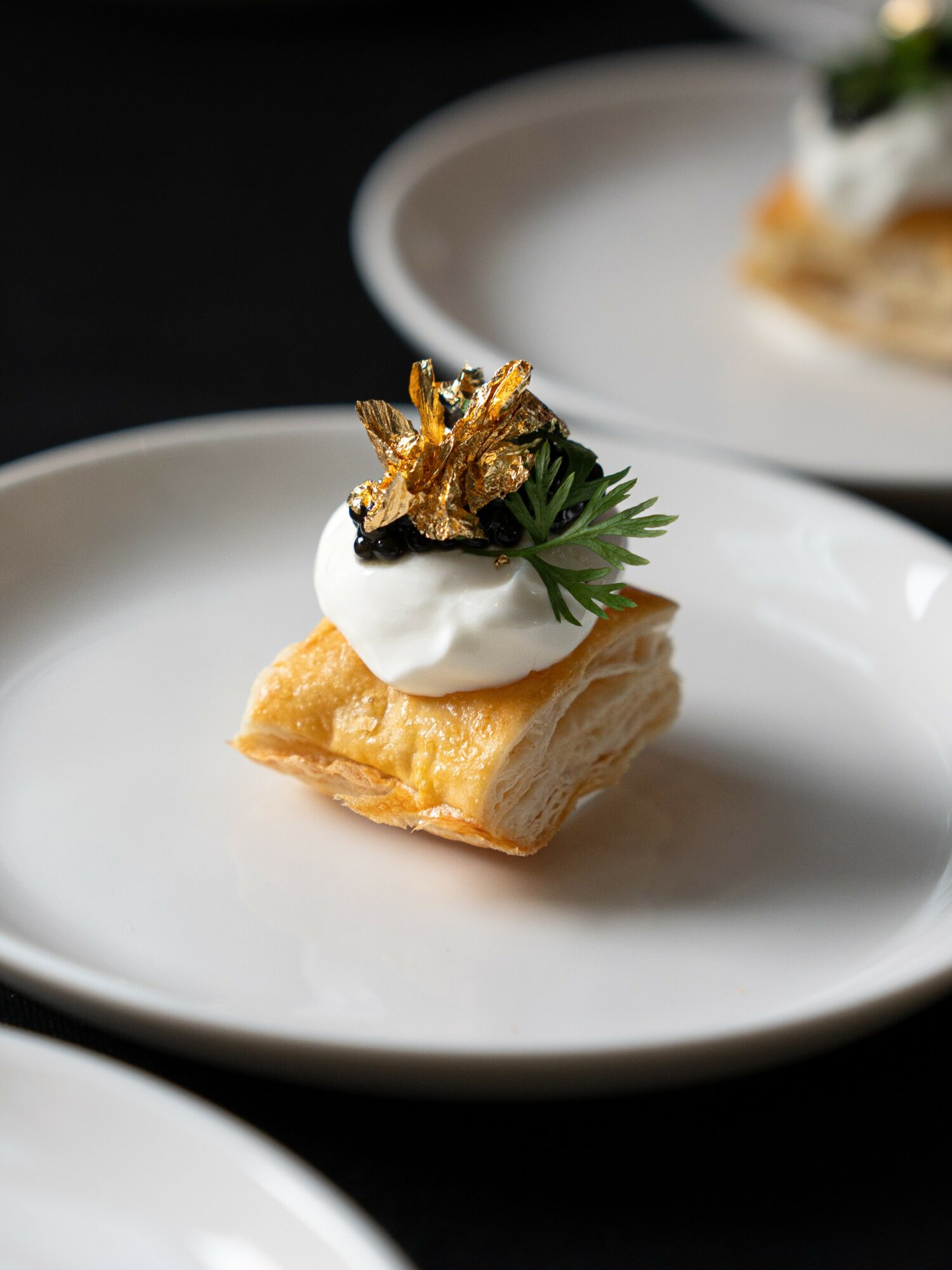 Pastry topped with whipped cream, gold leaf, and a small green herb on a white plate.
