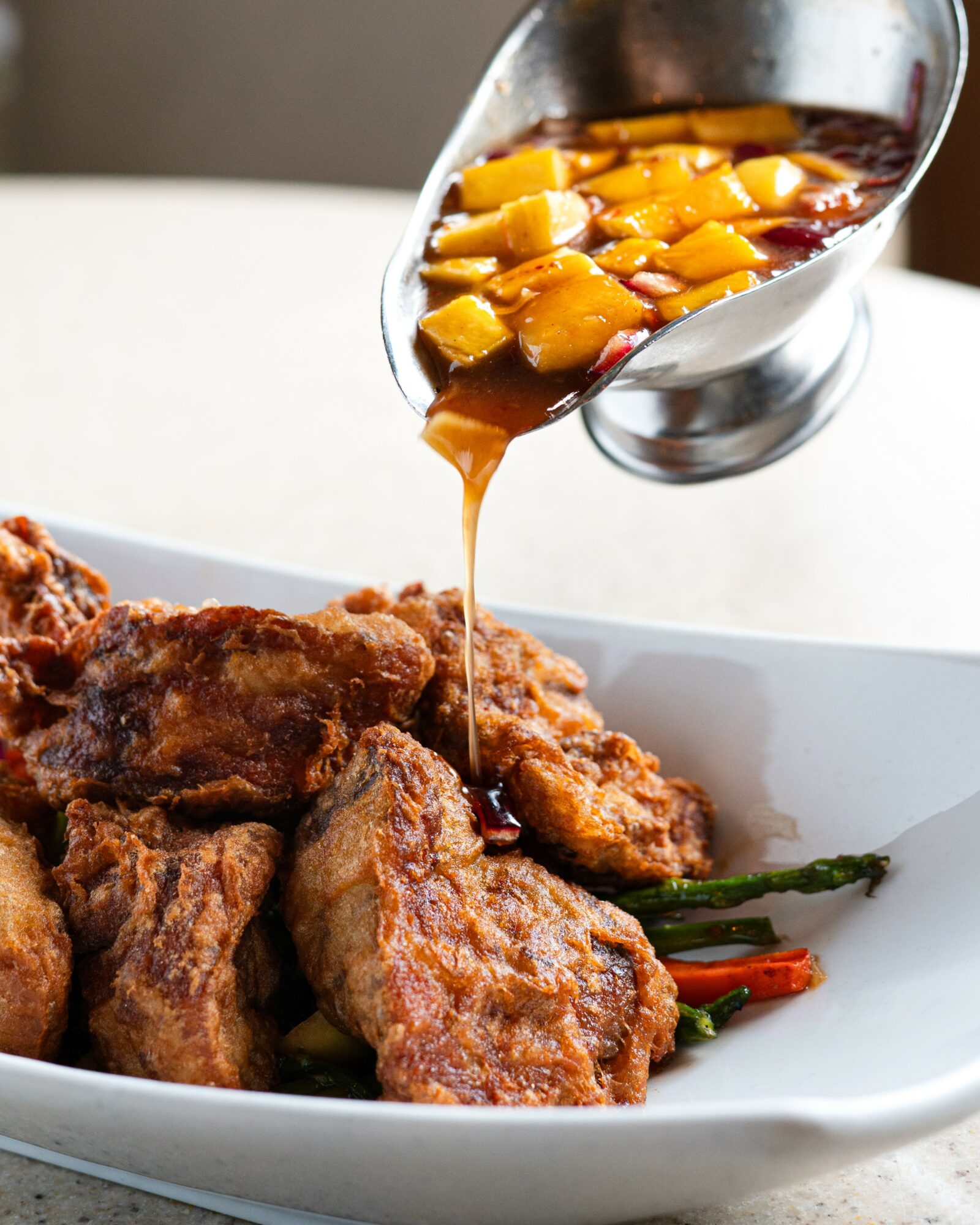 Fried chicken pieces with sauce being poured from a metal container, garnished with green beans and red peppers, in a white dish.