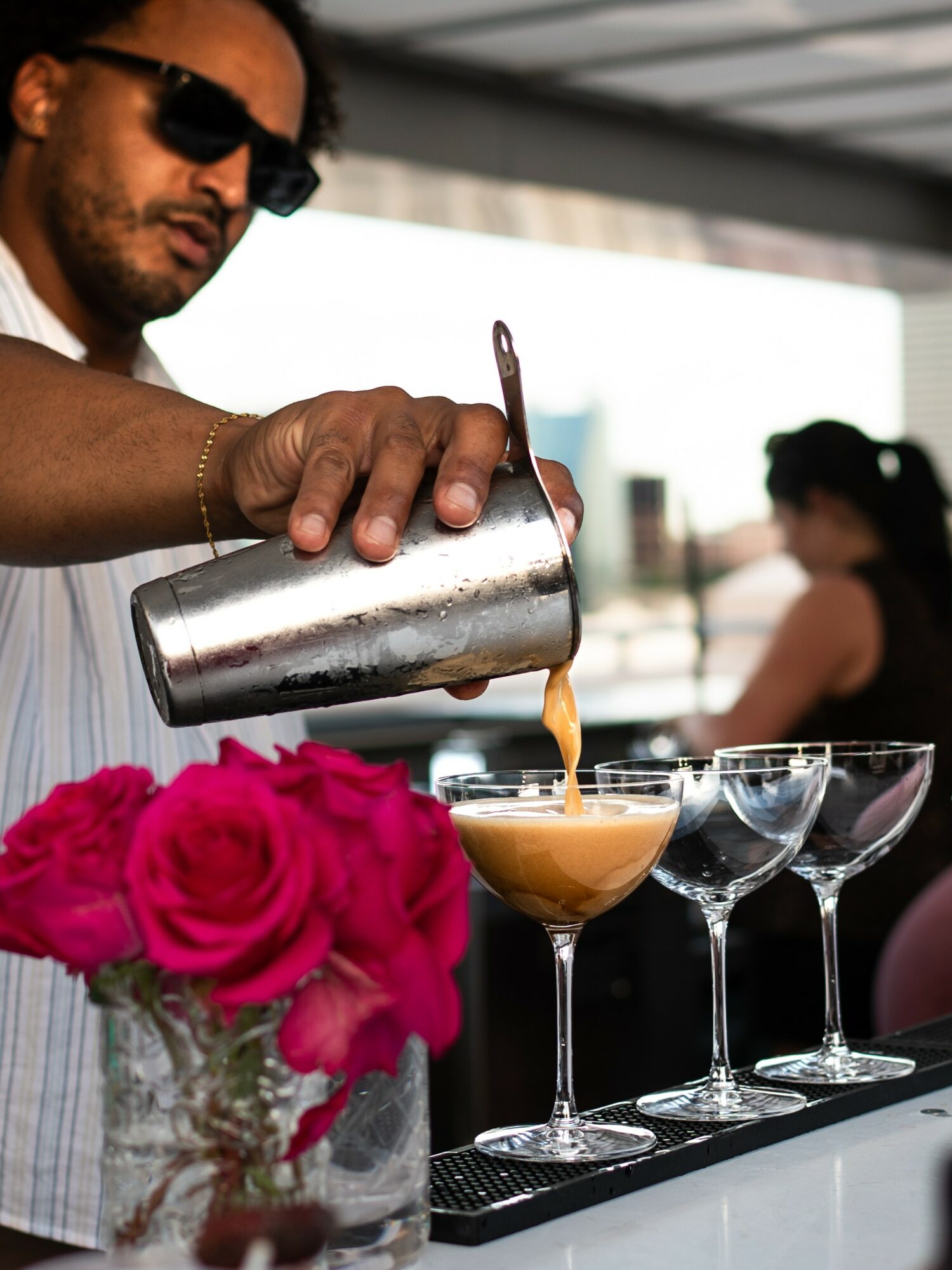 Barista pouring a drink into a glass with pink flowers in the foreground, and a woman in the background, in a cafe setting.