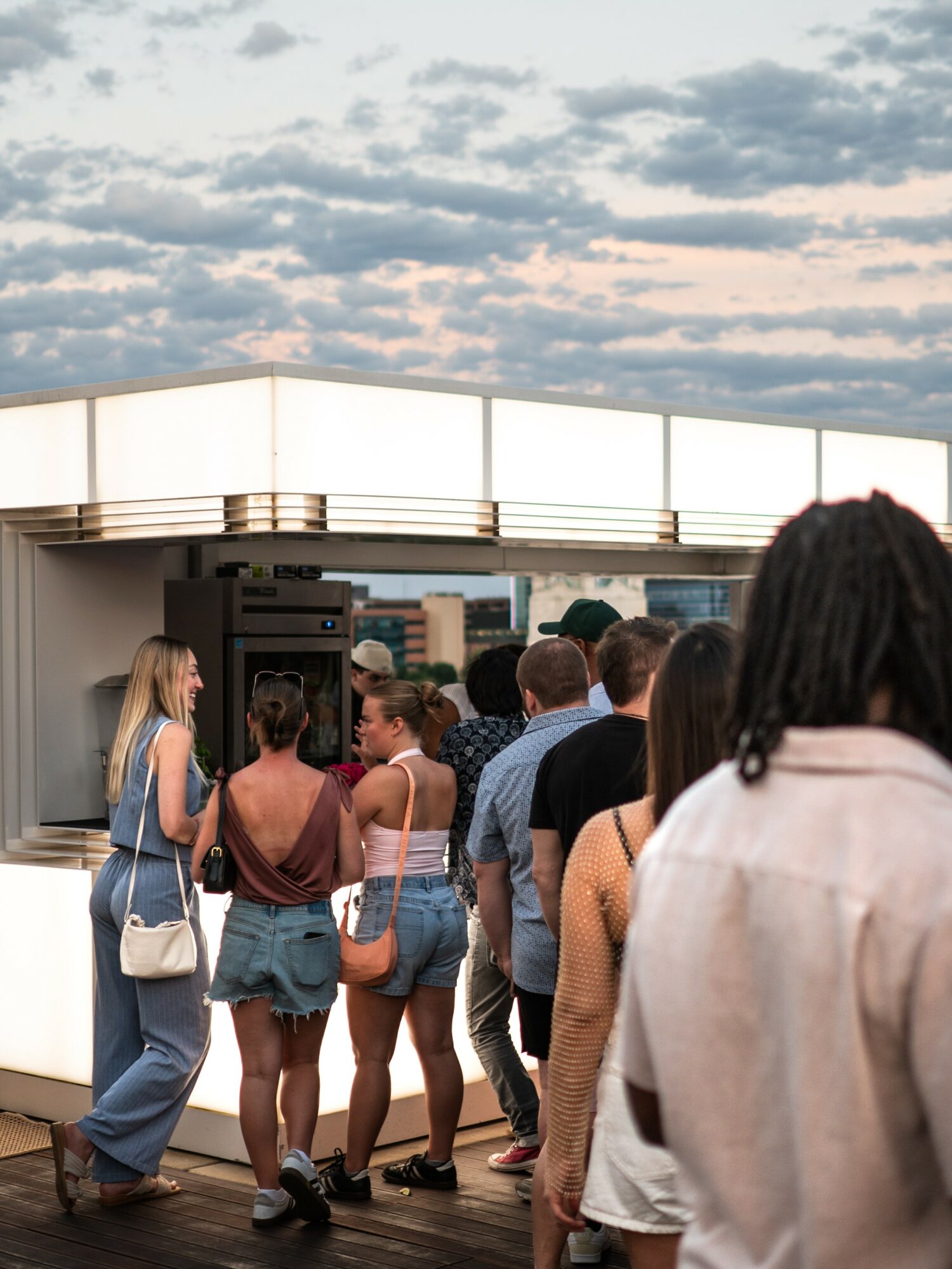 People standing in line outdoors under a cloudy sky, some with backpacks and casual clothing.