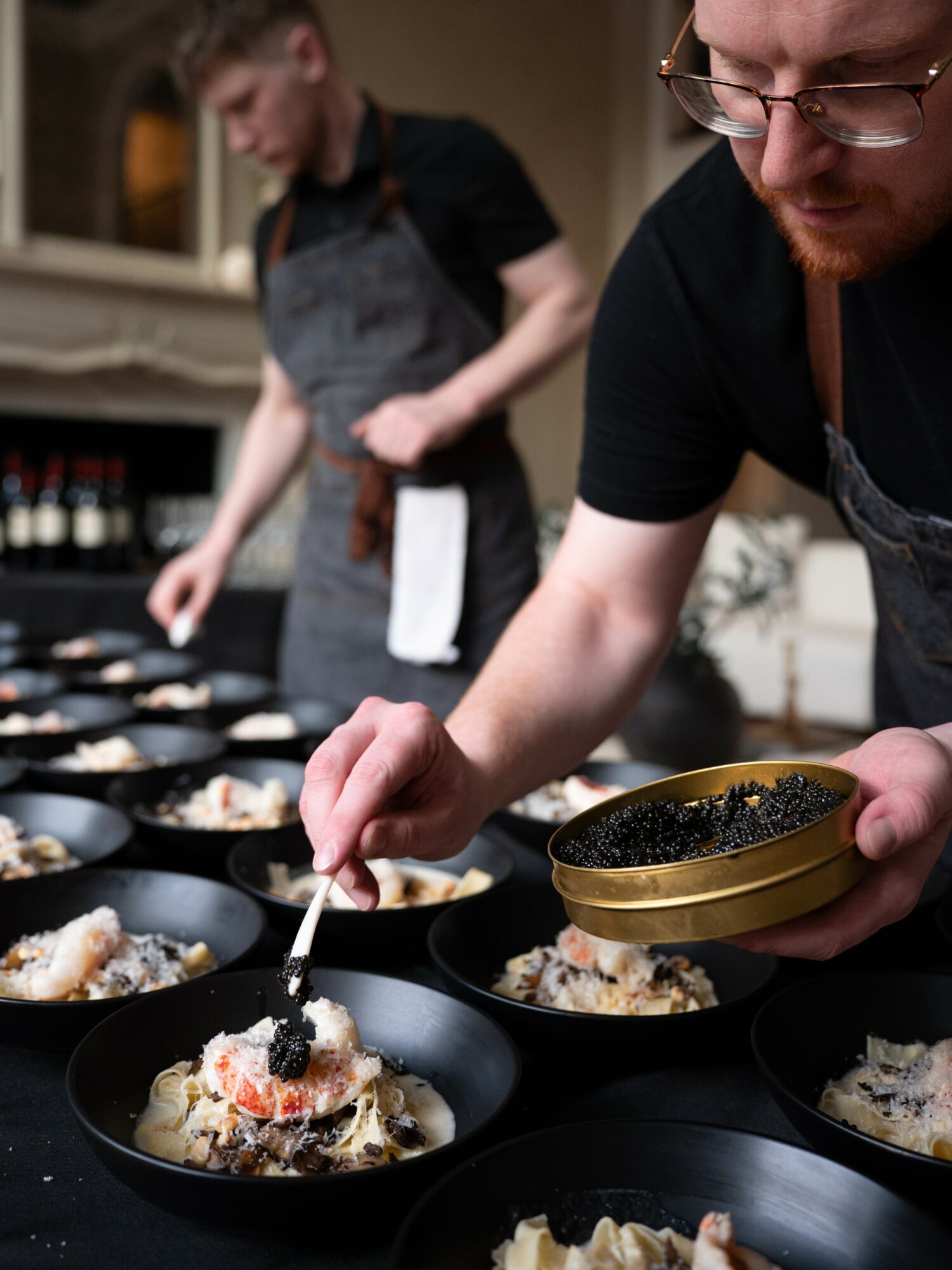 Two people preparing dishes in a kitchen, focusing on plating food in black bowls, with one person holding a bowl and spoon.