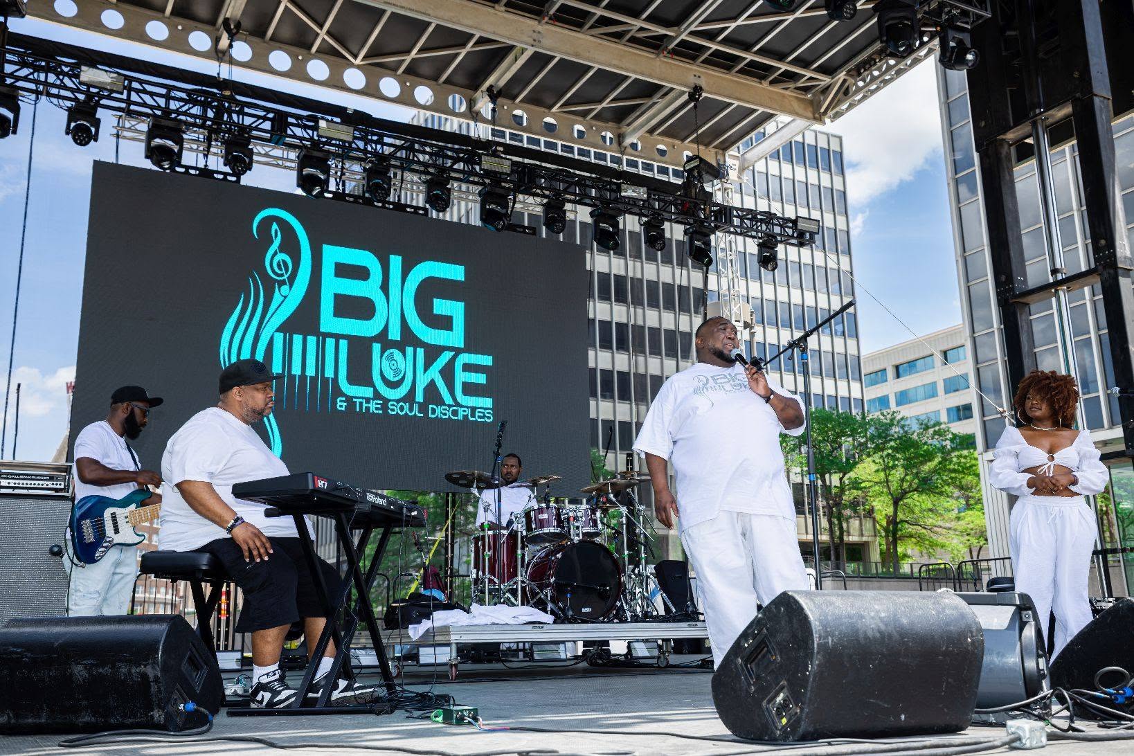 Musicians perform on an outdoor stage with a large screen displaying 'BIG LUKE & THE SOUL OSCILLATORS' behind them.