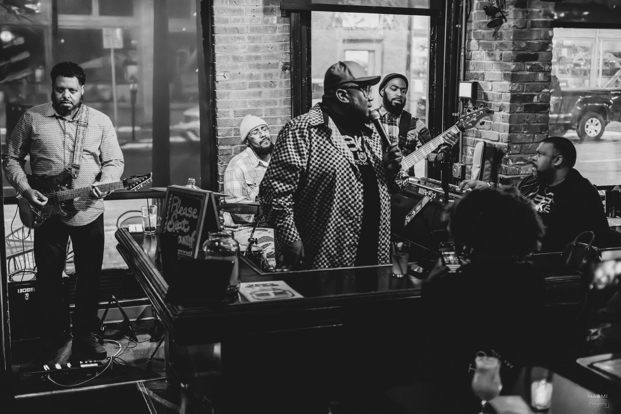 Group of musicians performing in a cafe with brick walls and large windows, playing guitars and singing.