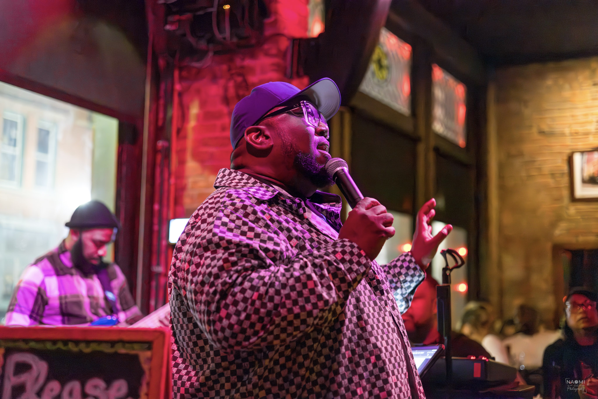 Person singing into a microphone in a lively indoor setting with colorful lighting and other people in the background.