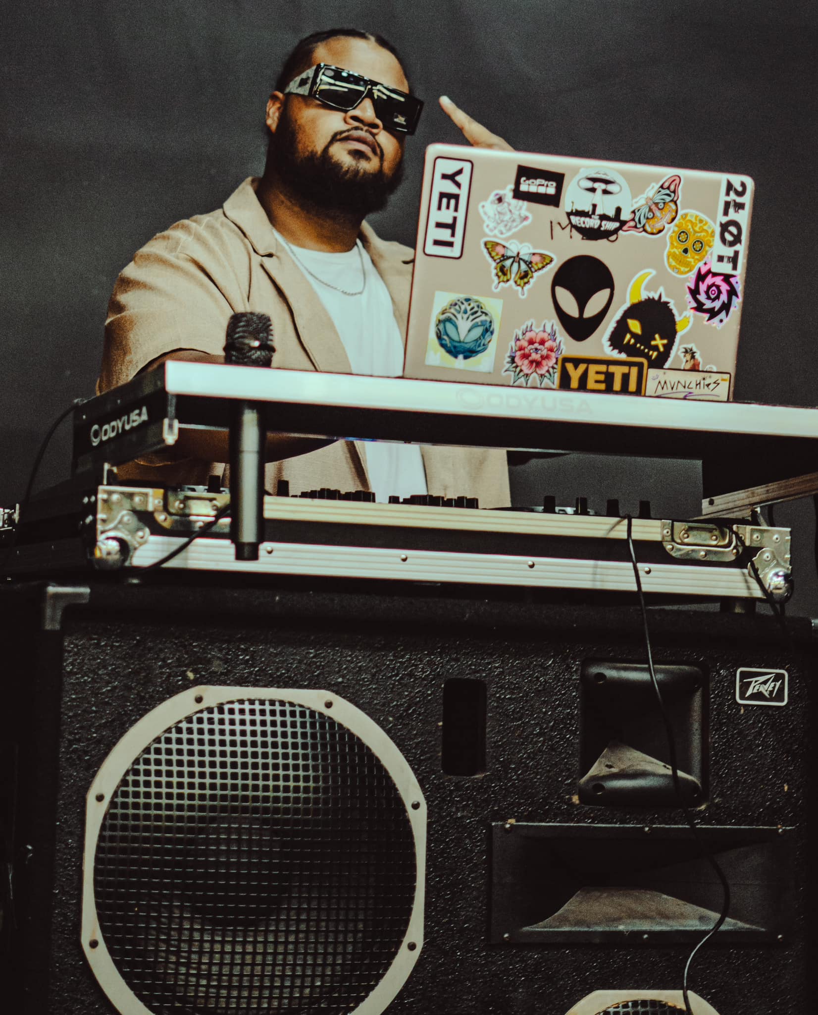 DJ with sunglasses and beard, standing behind a turntable, with a laptop covered in stickers, in front of a speaker.