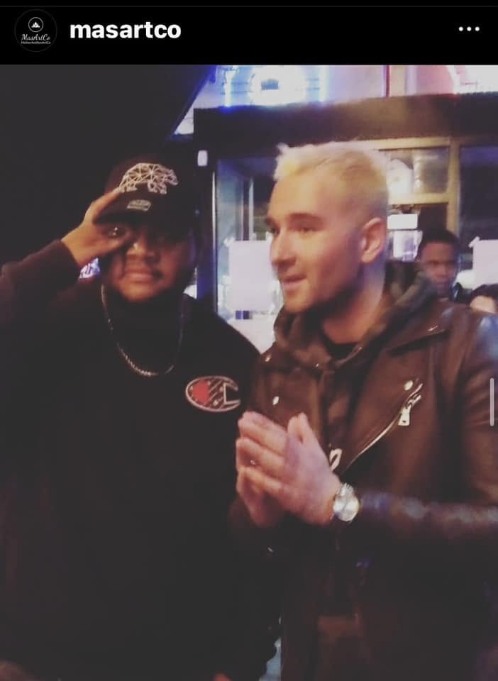 Two men standing together in a dimly lit setting, one wearing a black cap and jacket, the other with platinum blonde hair and a brown leather jacket.