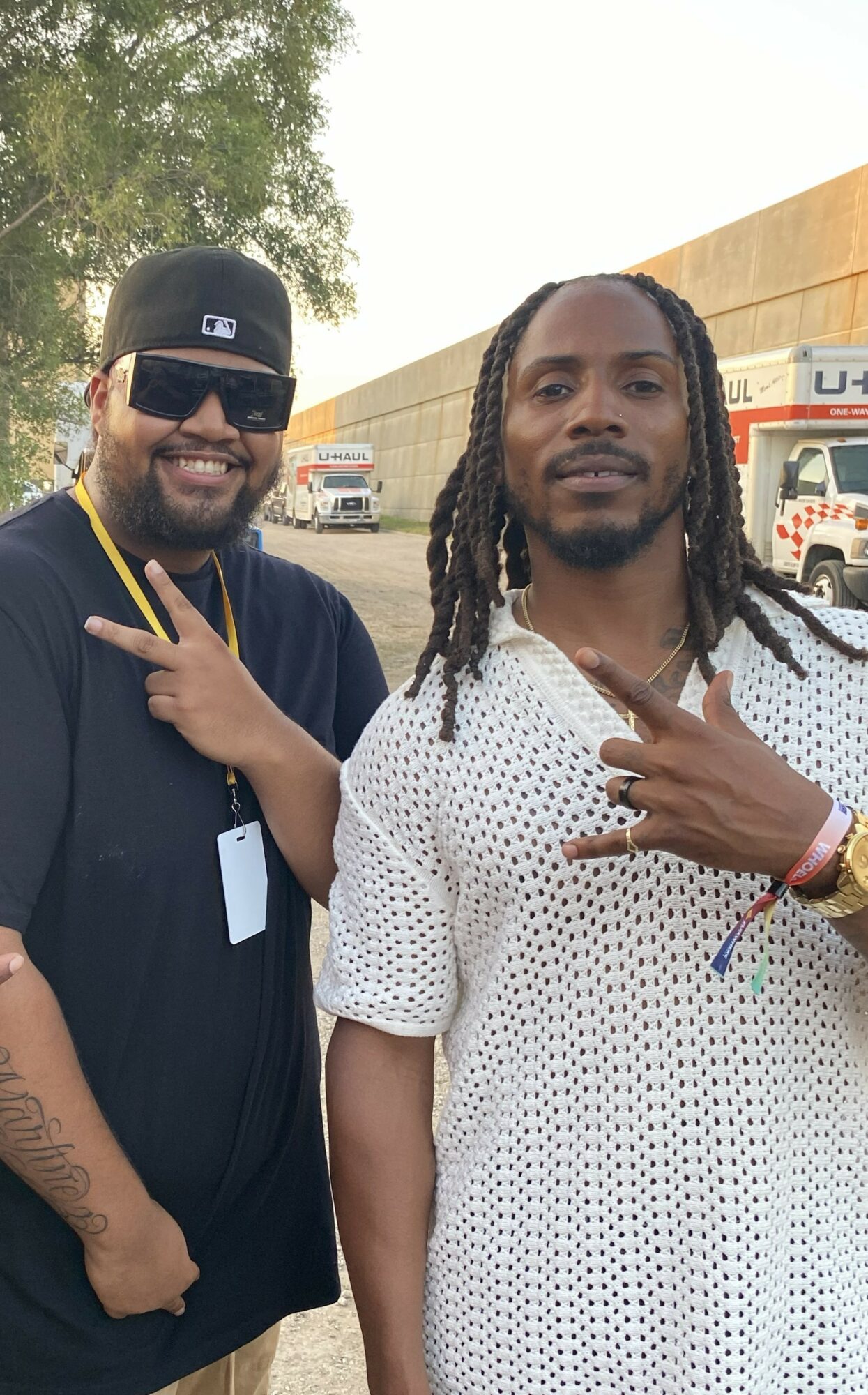 Two men standing outdoors, one wearing sunglasses and a cap, the other with long dreadlocks and a patterned shirt, both making hand gestures.