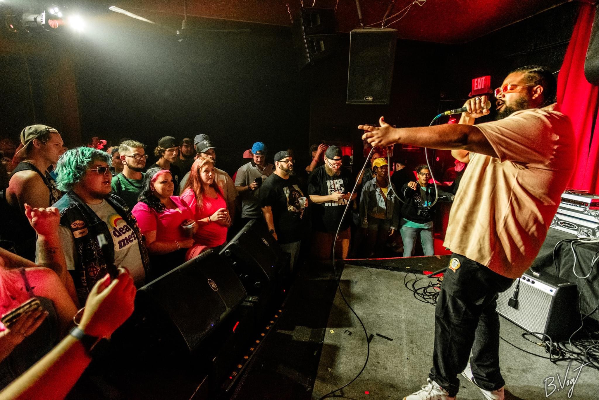A performer on stage gestures towards an audience in a dimly lit venue with red curtains and sound equipment.