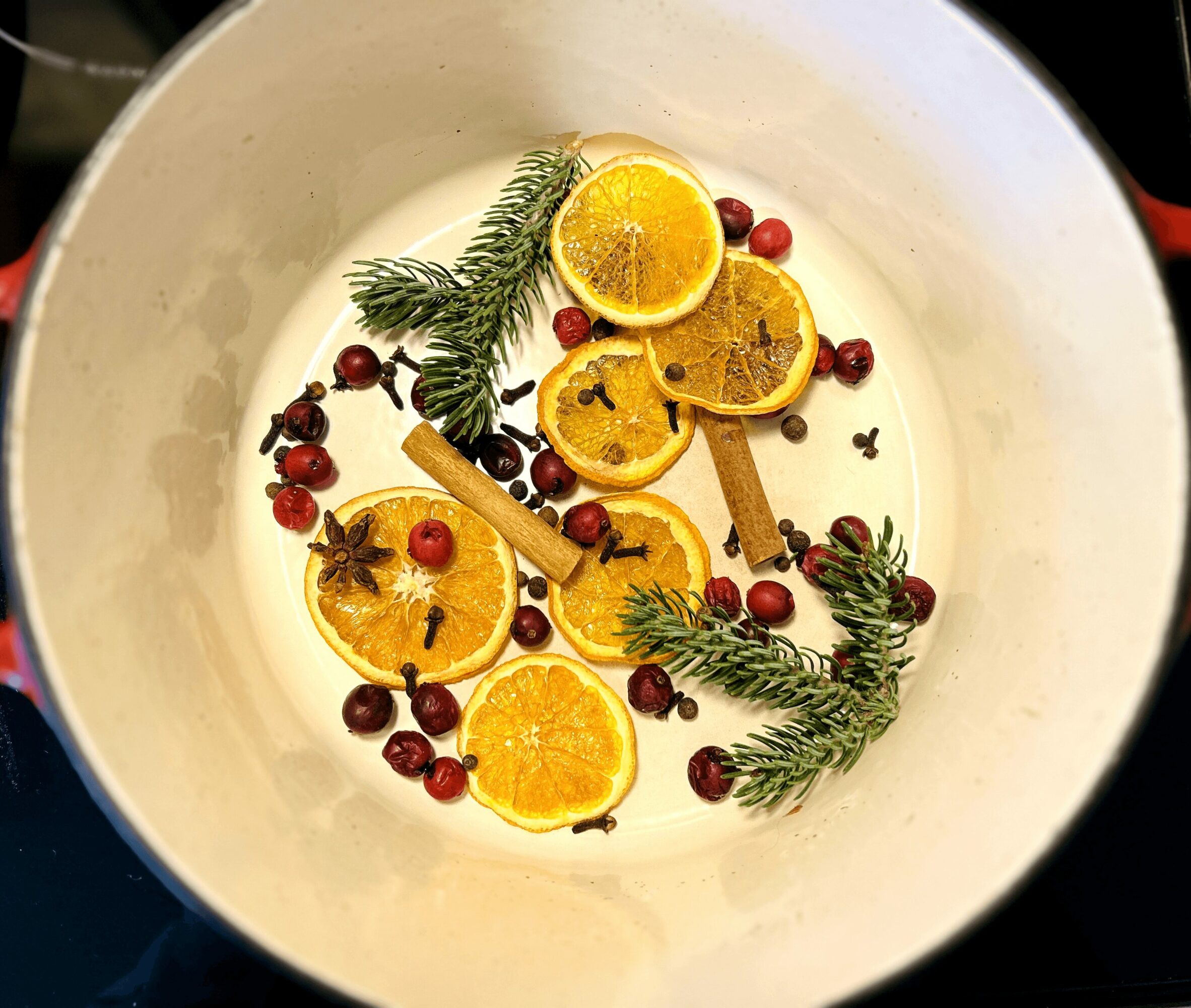 A bowl with slices of orange, cinnamon sticks, pine branches, and red berries in a creamy liquid.