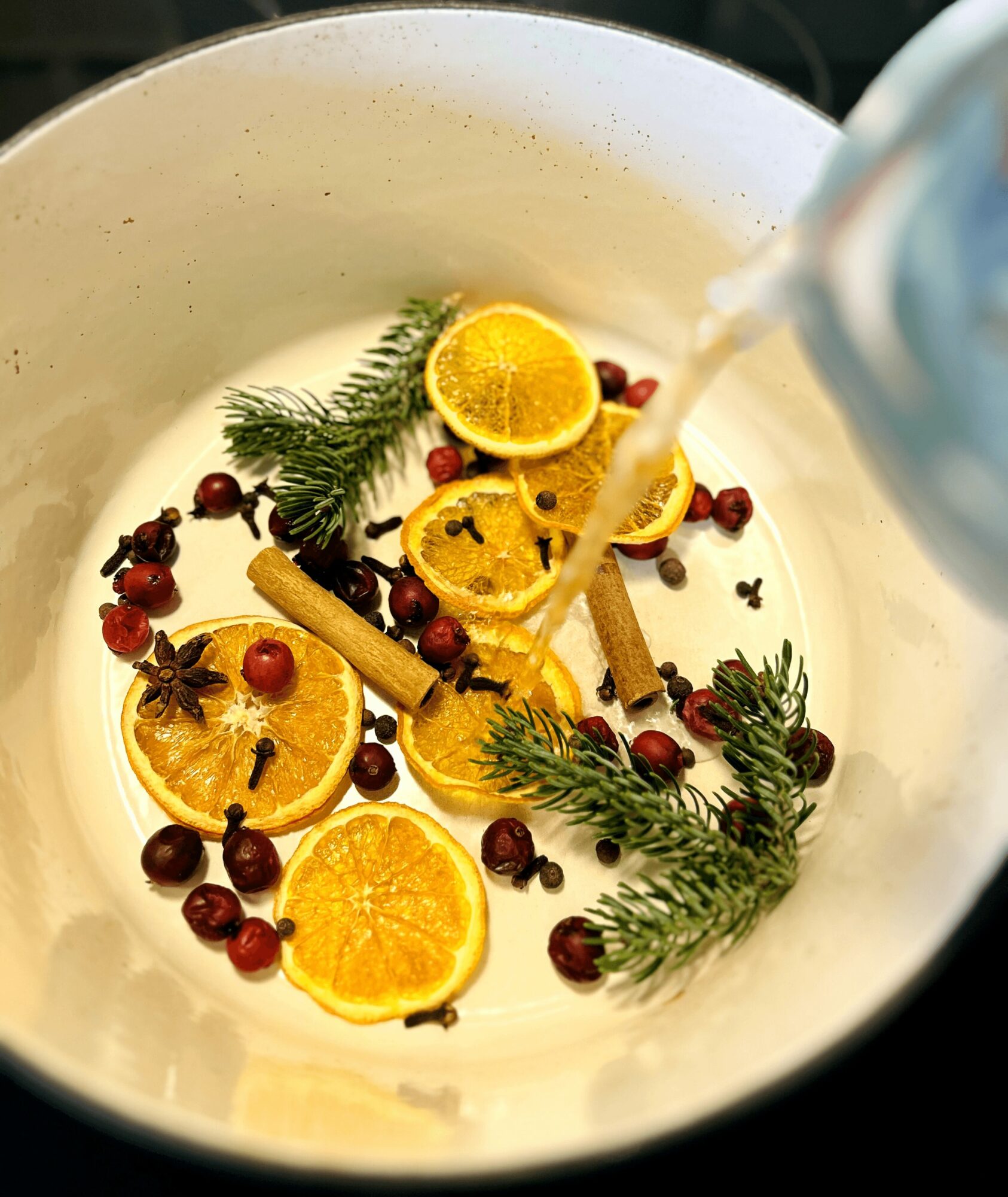 A white bowl with orange slices, cinnamon sticks, red berries, and green pine branches, with liquid being poured in.