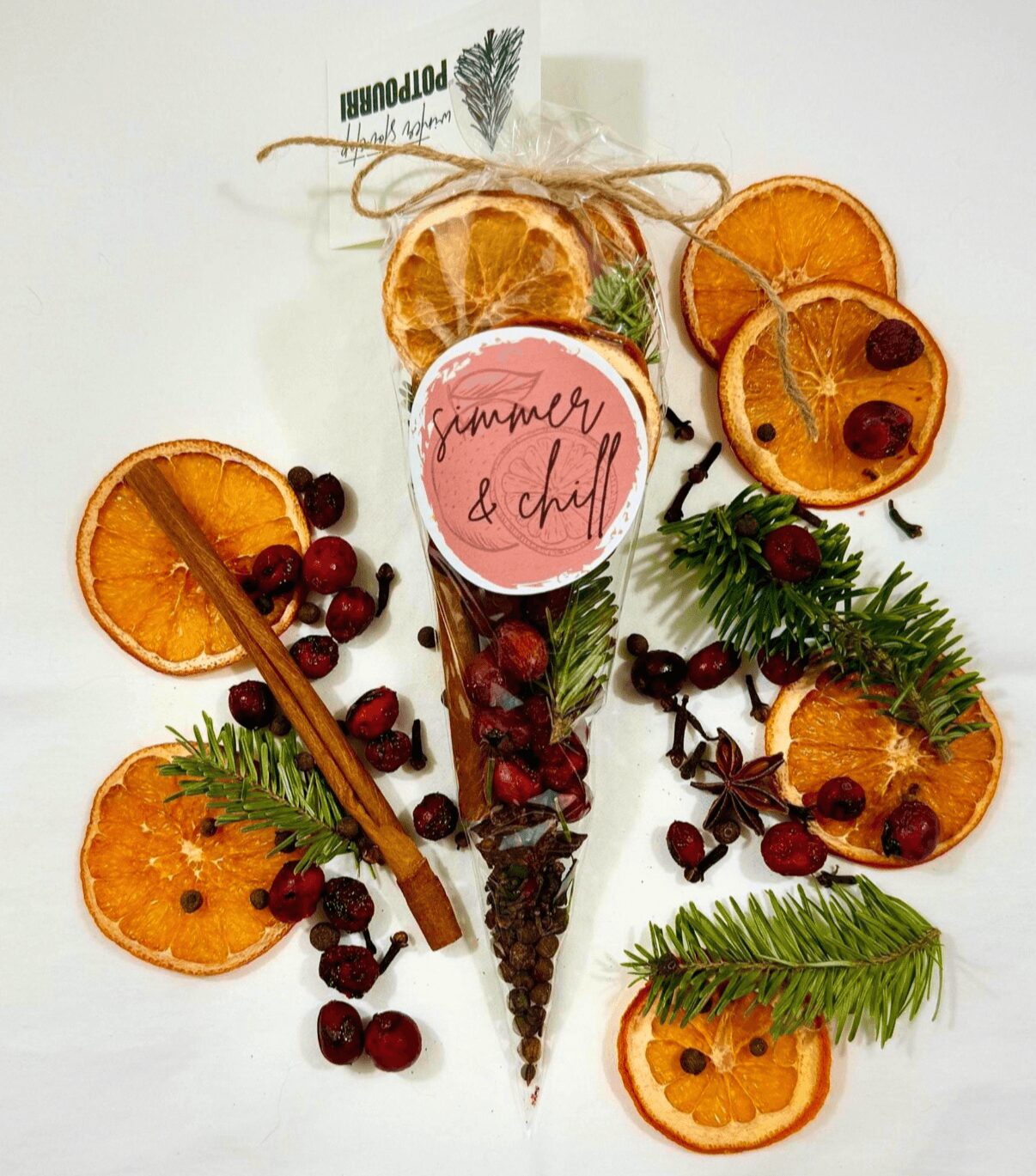 Decorative arrangement of dried orange slices, cherries, cinnamon stick, and green leaves on a white background with a pink circular label.