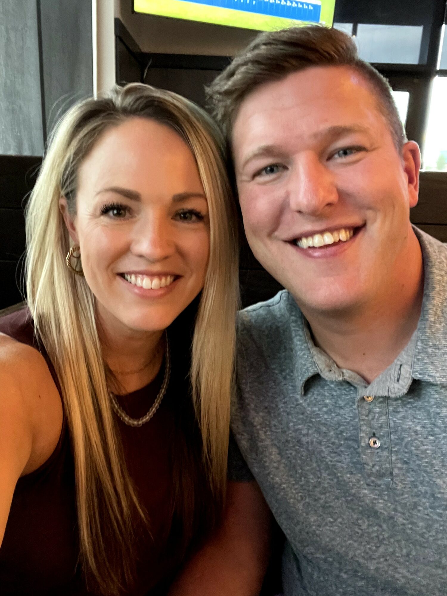 Two smiling people, a woman with long blonde hair and a man with short brown hair, pose together indoors.
