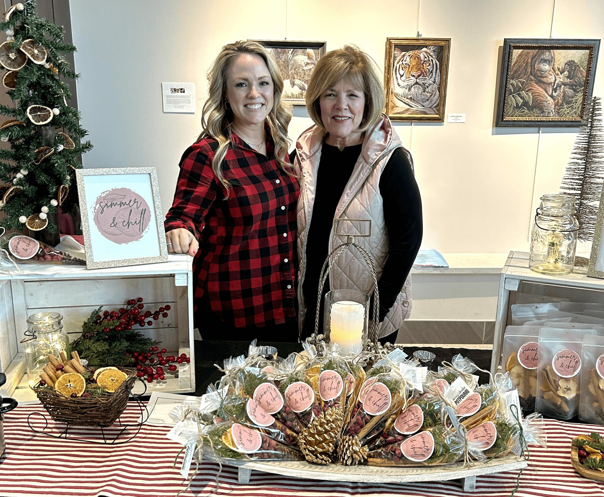 Two women stand behind a table with decorated pinecones and pink macarons, in a festive indoor setting with artwork on the wall.