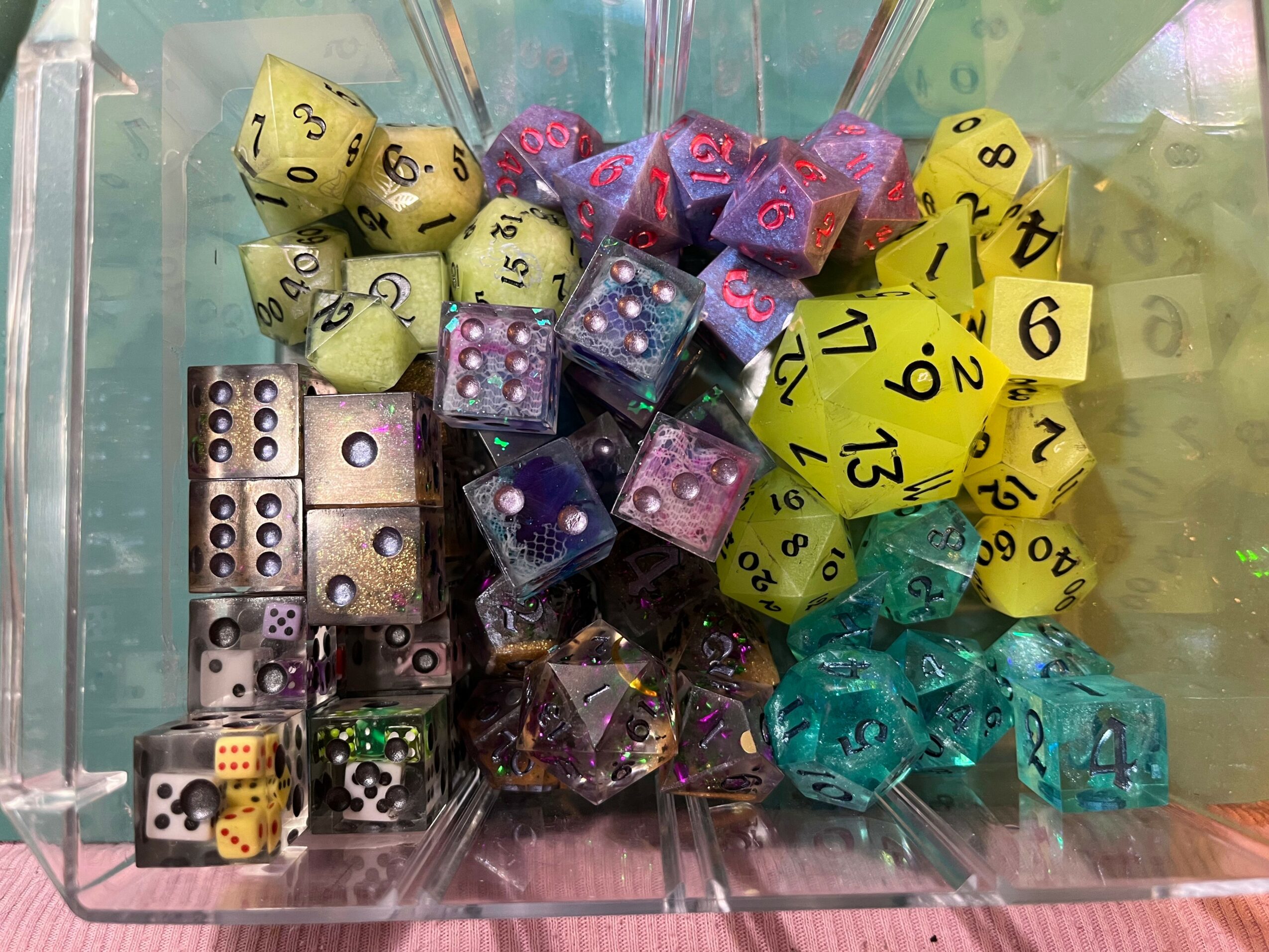 Container with colorful dice, including yellow, purple, blue, and green, some with numbers visible, in a transparent box.