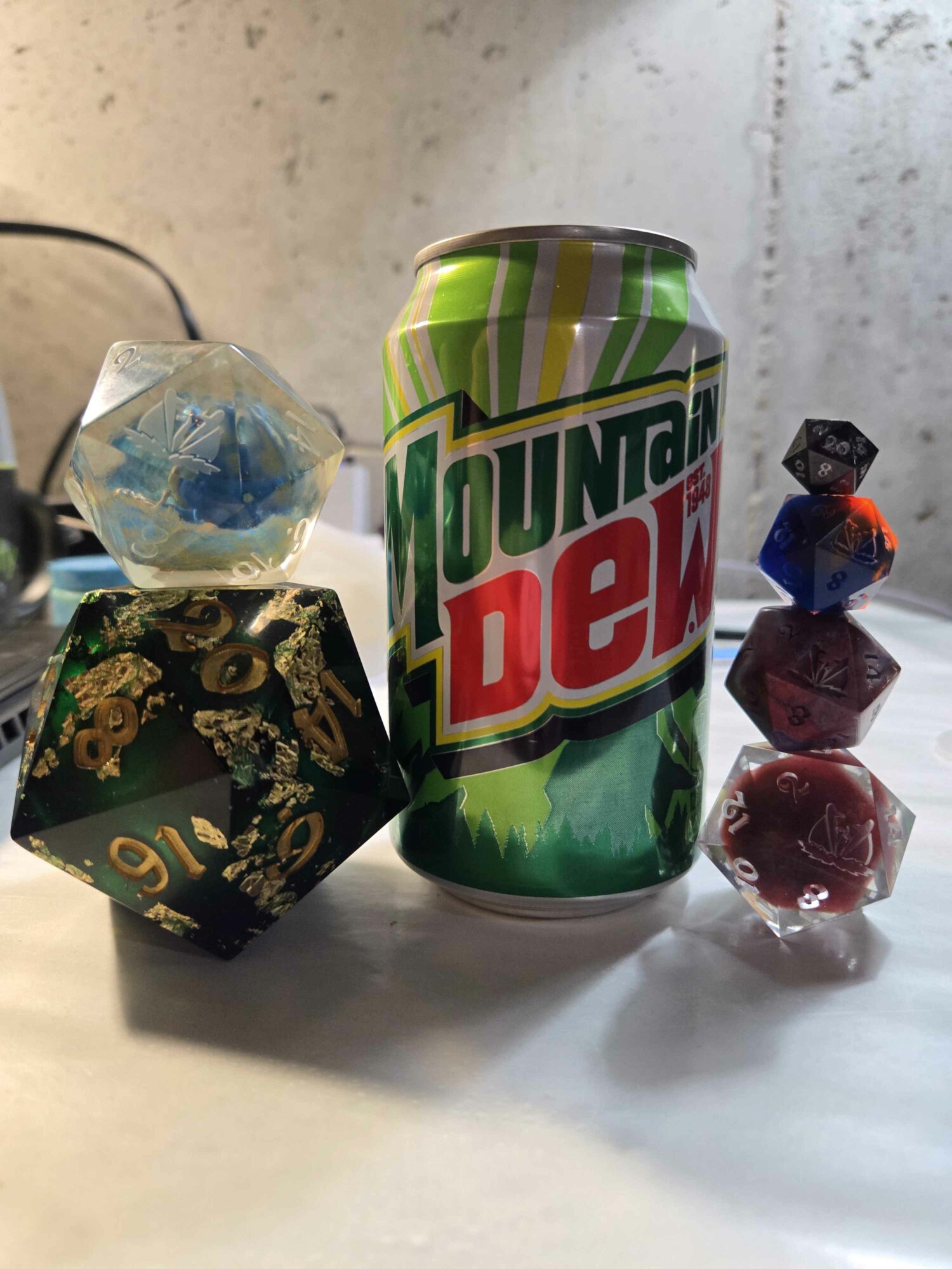A can of Mountain Dew, a decorative black and gold die, and a small stack of colorful dice on a white surface.