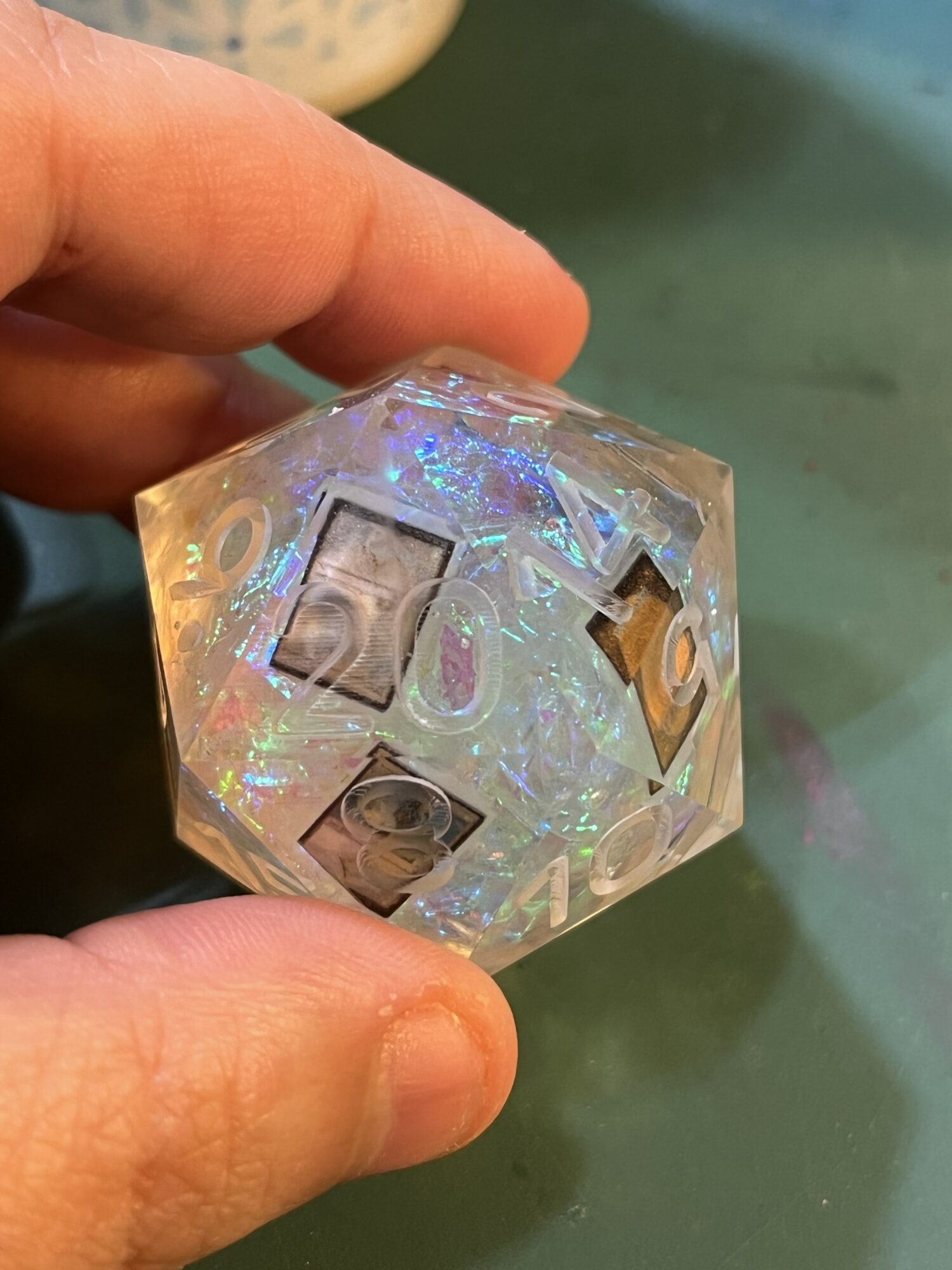 Hand holding a transparent, holographic, 20-sided die with numbers visible, against a green background.
