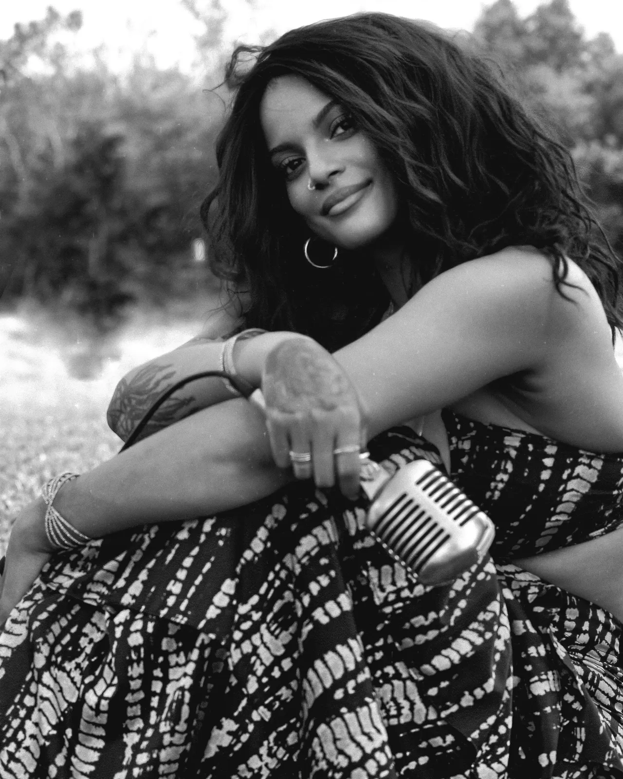 Woman with curly hair sitting outdoors, holding a vintage camera, smiling, in black and white.
