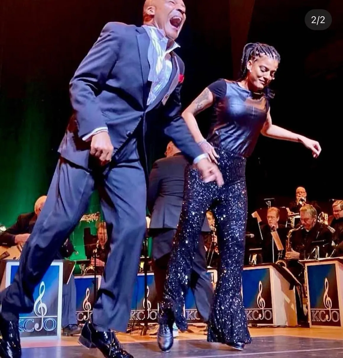 Two performers dancing on stage with an orchestra in the background, one man in a suit and one woman in sparkly pants.