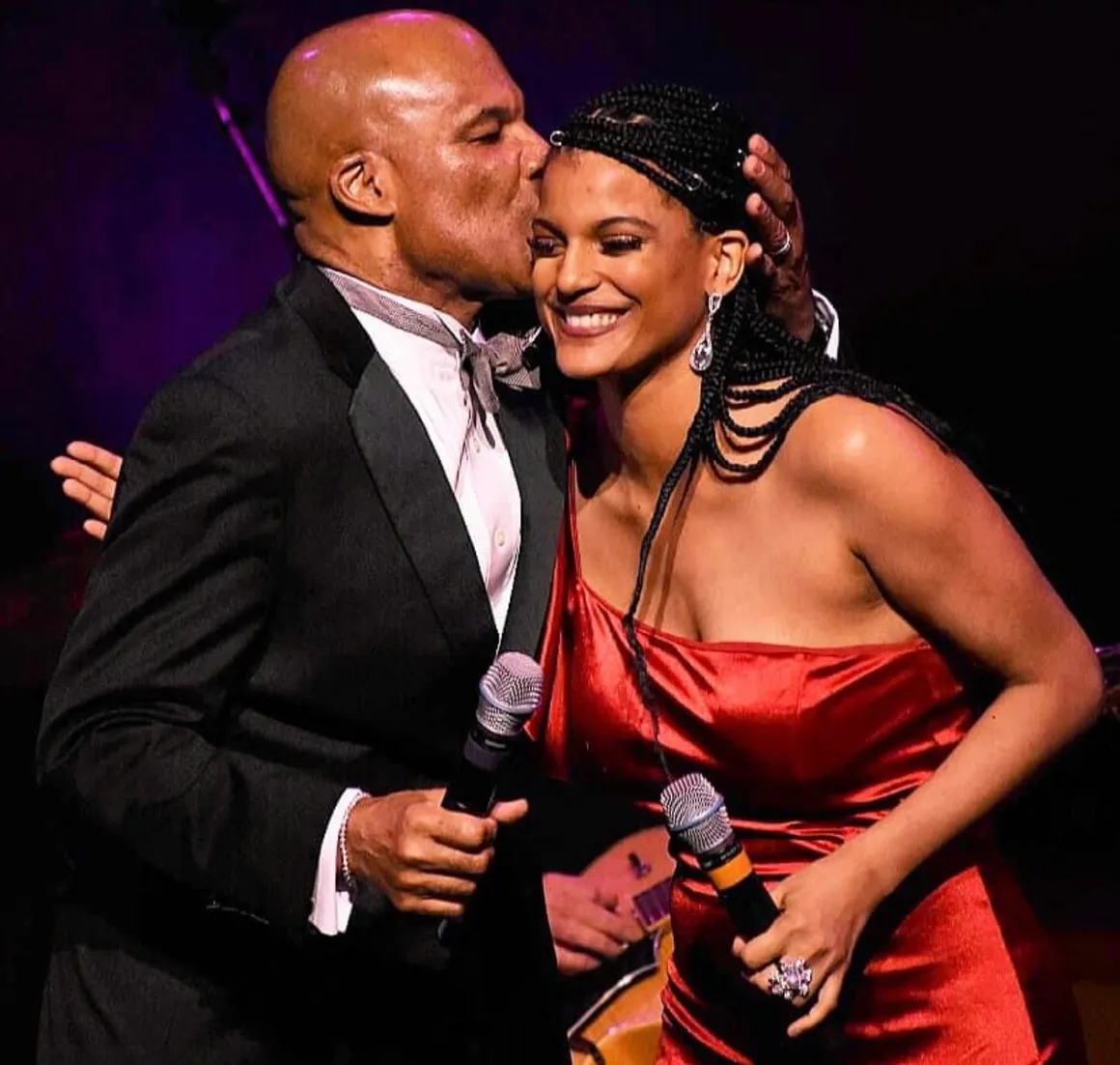 Two people smiling, one kissing the other's cheek, holding microphones, in a dark setting with purple lighting.